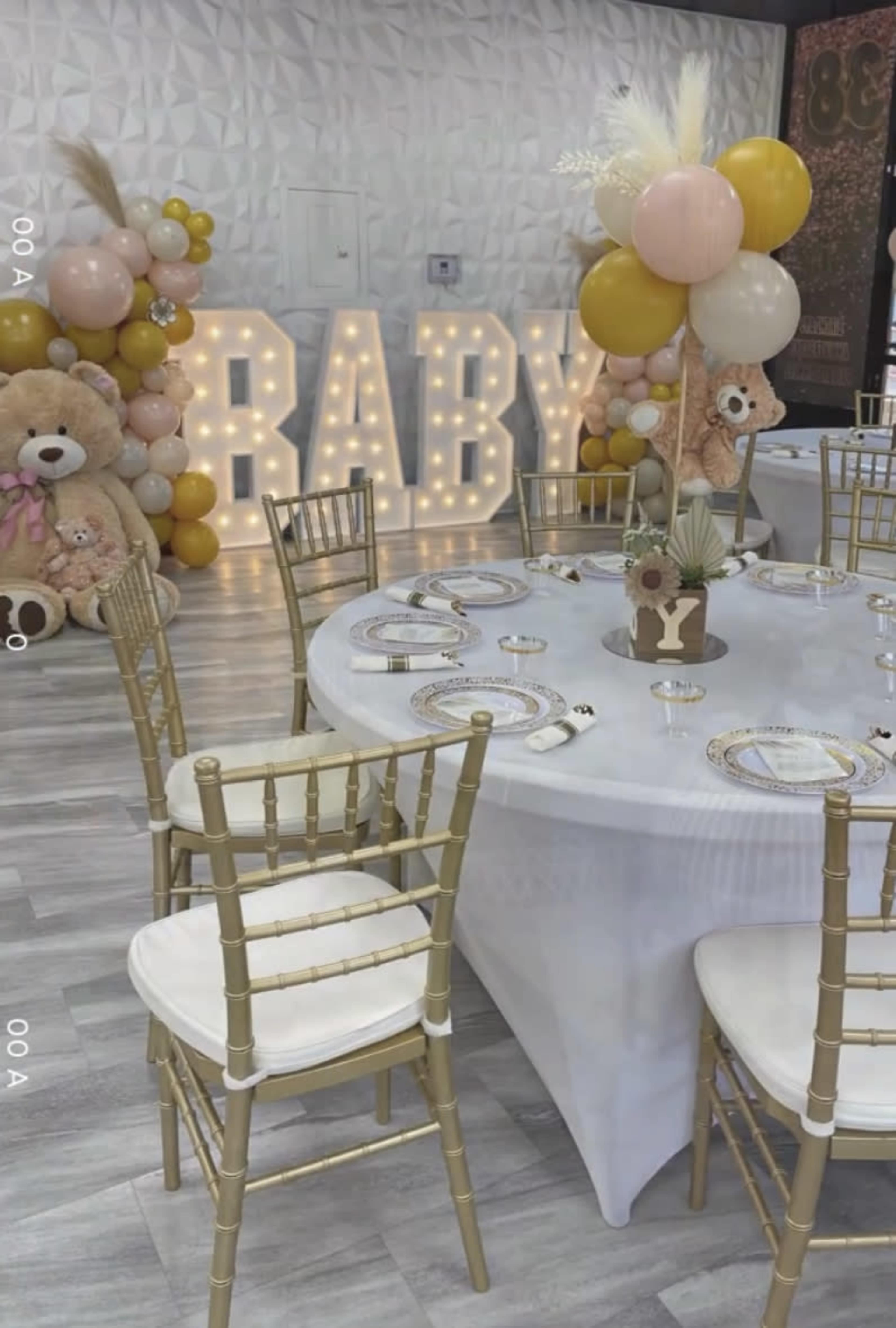 A decorated event space for a baby celebration, featuring a large illuminated "BABY" sign, colorful balloon arrangements, and a round table set with gold chairs.
