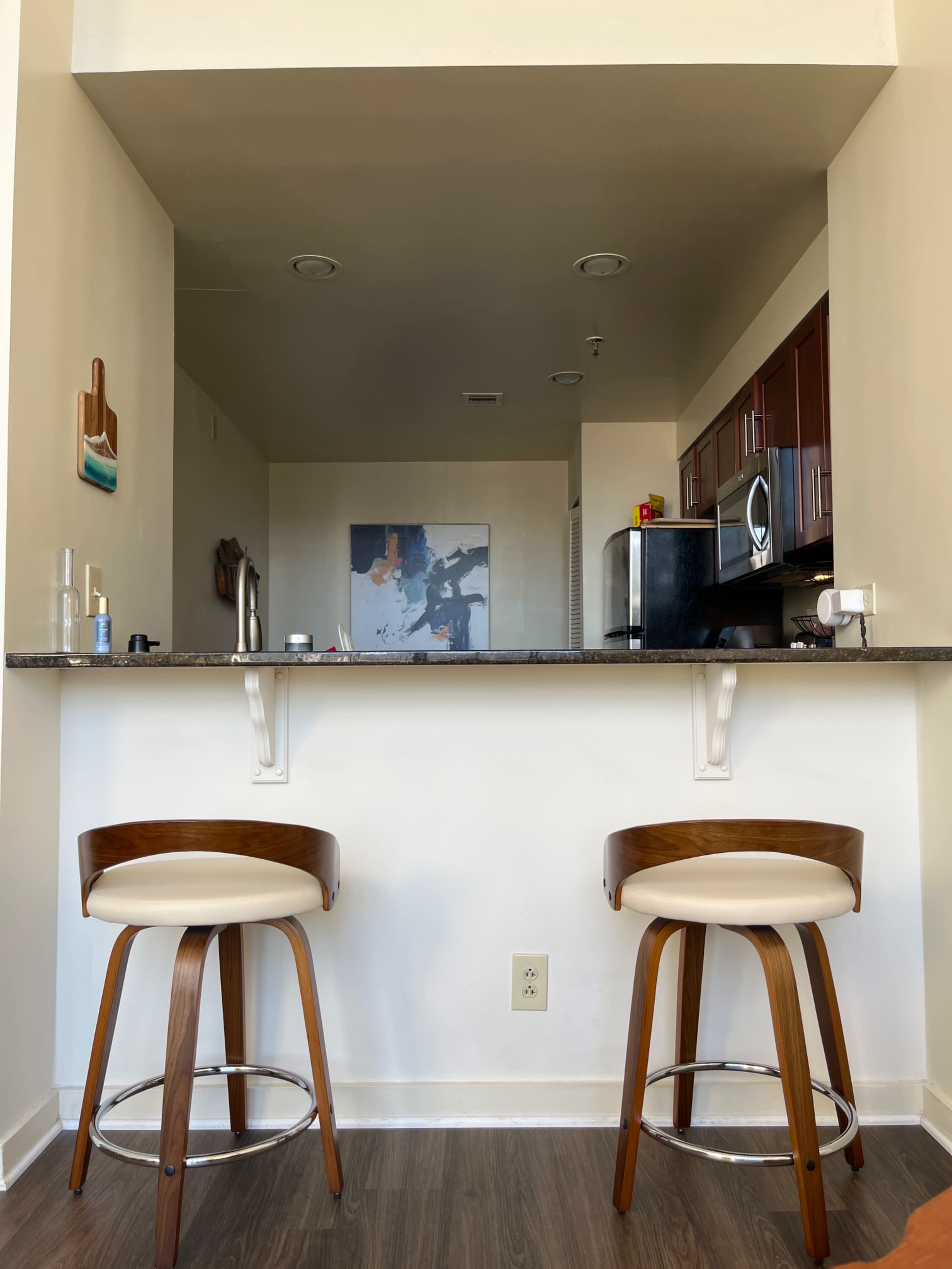 The image shows a modern kitchen bar area with two wooden stools and a view of the kitchen behind a granite countertop.