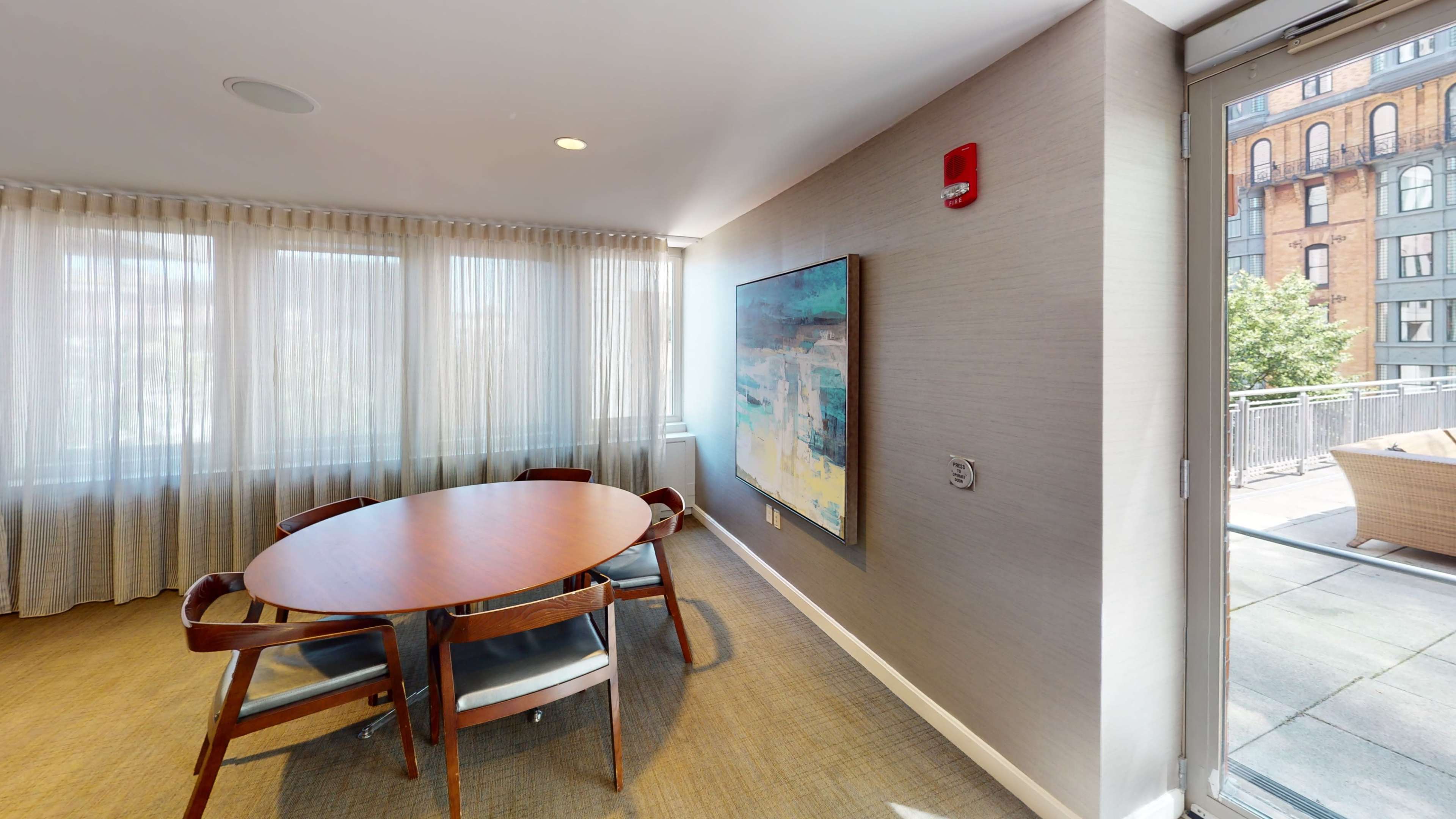 The image shows a conference room with a round wooden table, surrounded by chairs, and large windows displaying light-colored curtains.