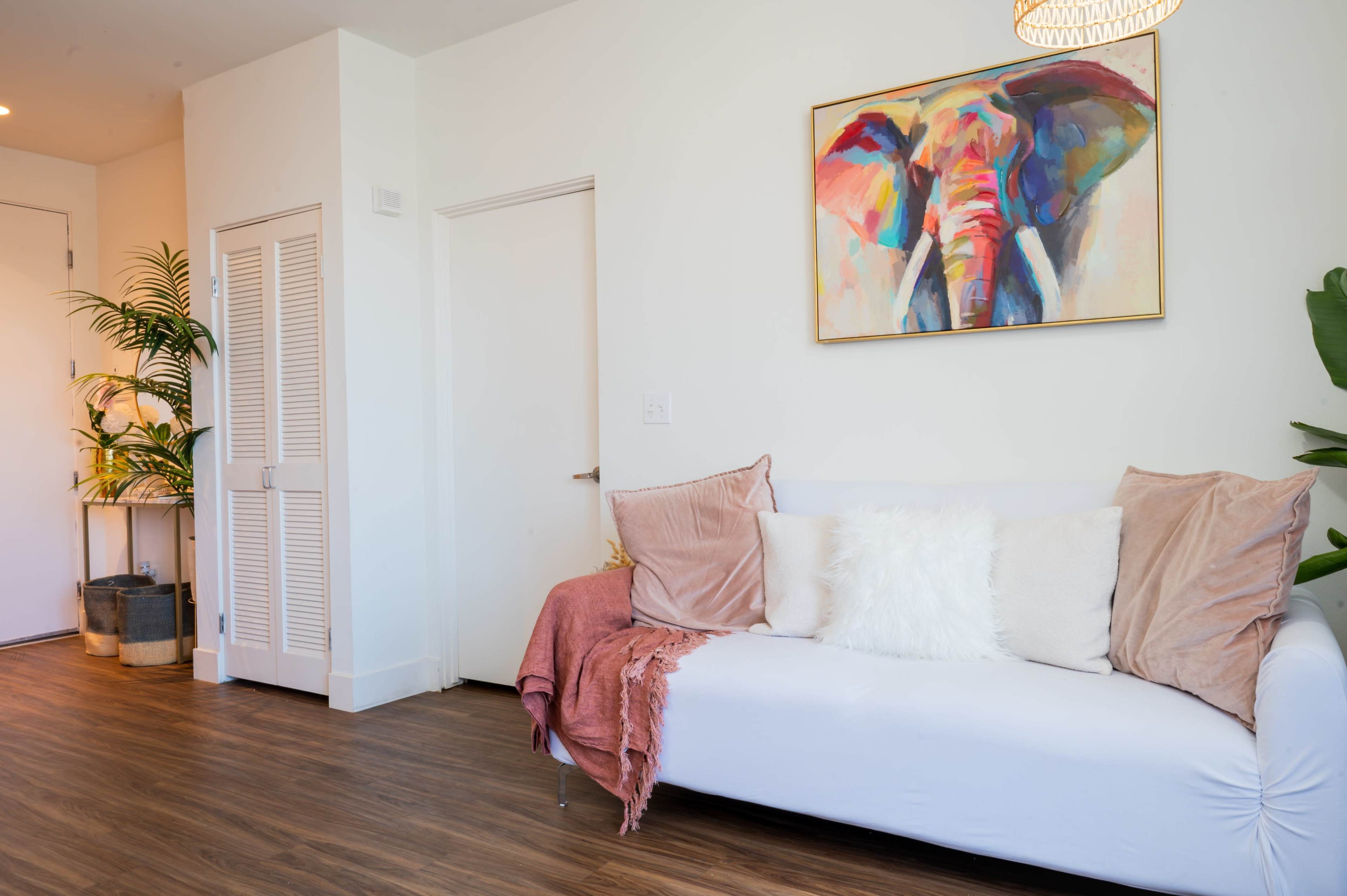 The image shows a modern living room featuring a white sofa adorned with pillows and a blanket, an elephant painting on the wall, and greenery in the corners.