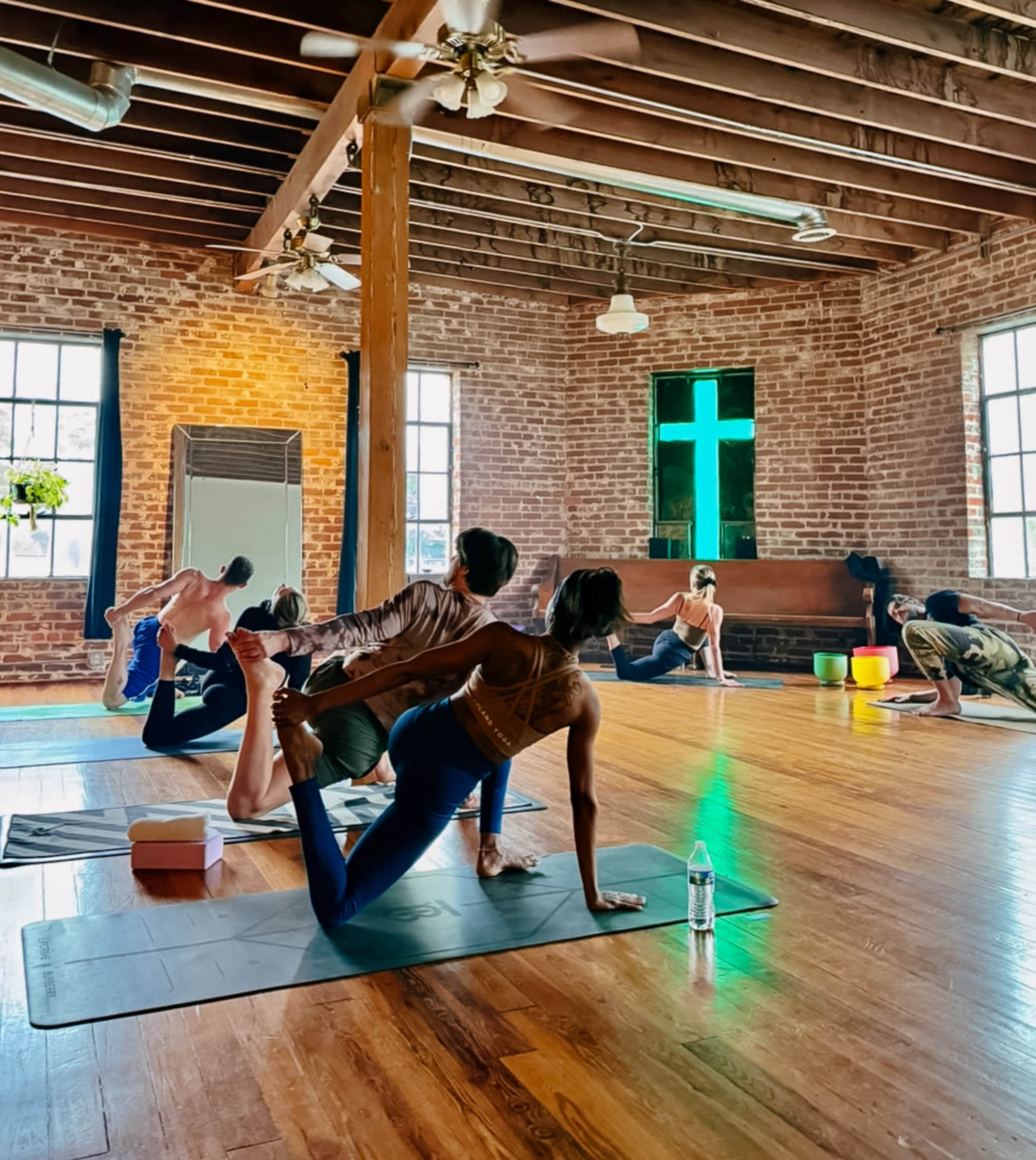 A group of people are practicing yoga on mats in a spacious room with exposed brick walls and large windows.