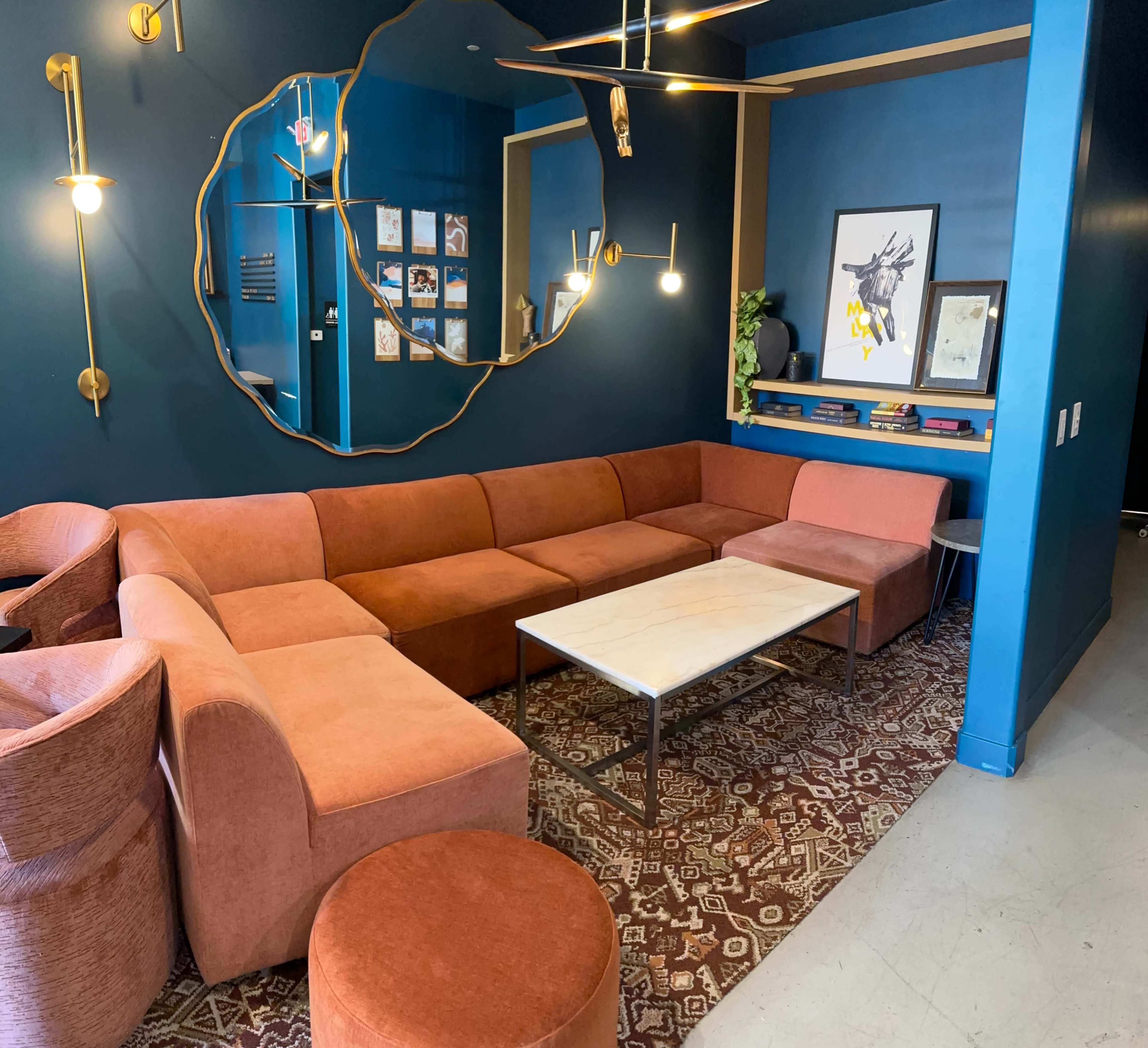 The image shows a modern lounge area with an L-shaped orange couch, a marble coffee table, and decorative elements on a deep blue wall.