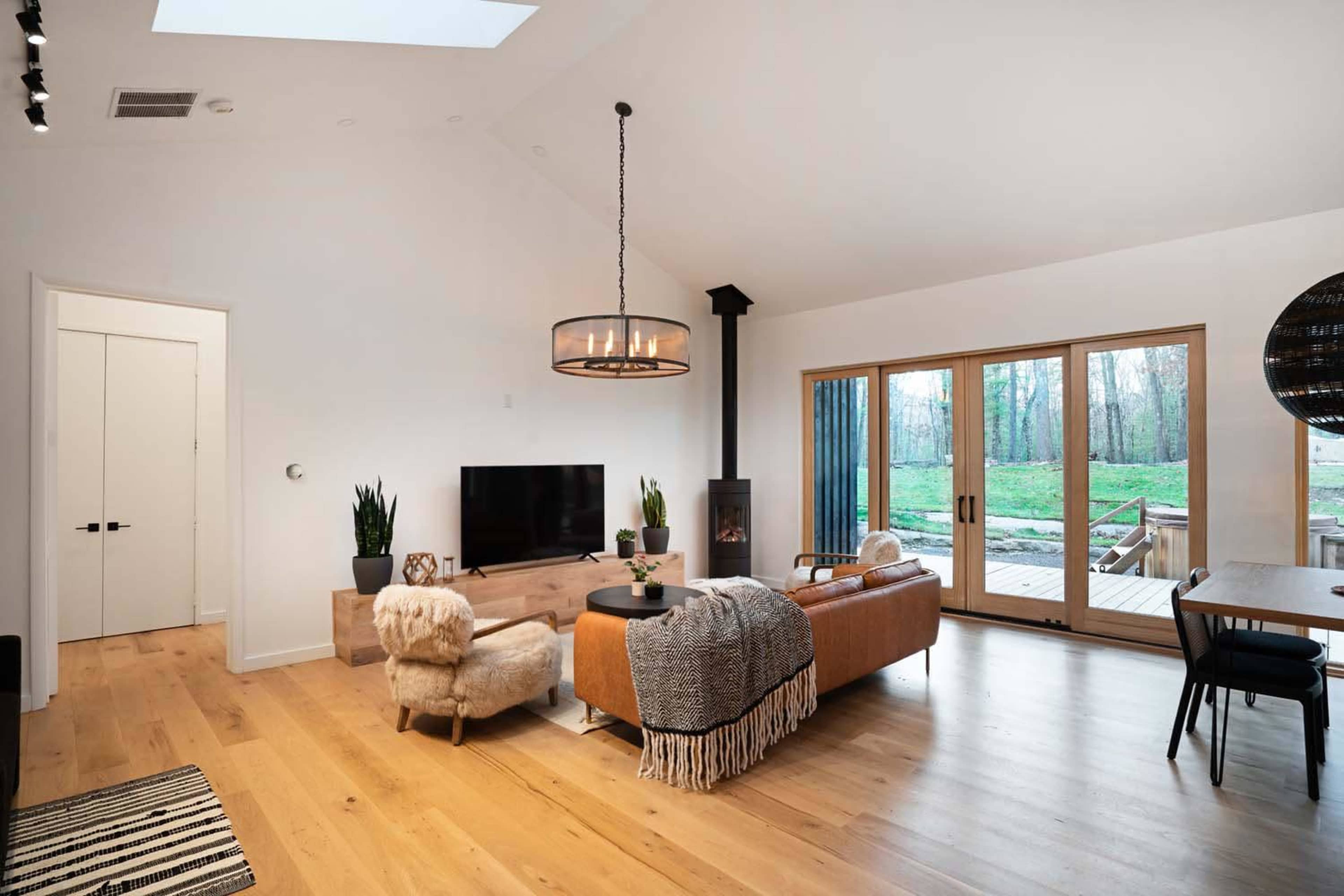 The living room features a modern design with a large window overlooking a deck and green outdoor space, a television on a wooden console, and a cozy seating area.