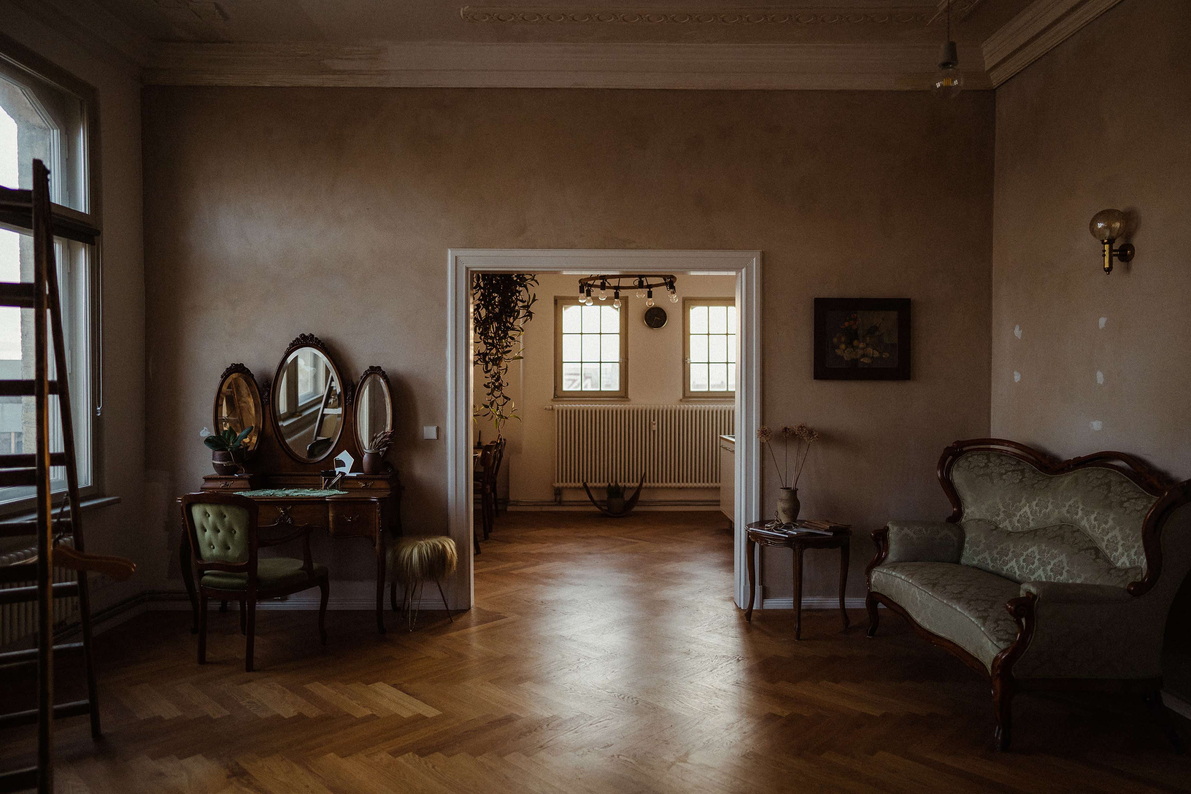 A spacious, softly lit room with a vintage vanity table, a green upholstered chair, and a patterned sofa, leading to another room through an arched doorway.