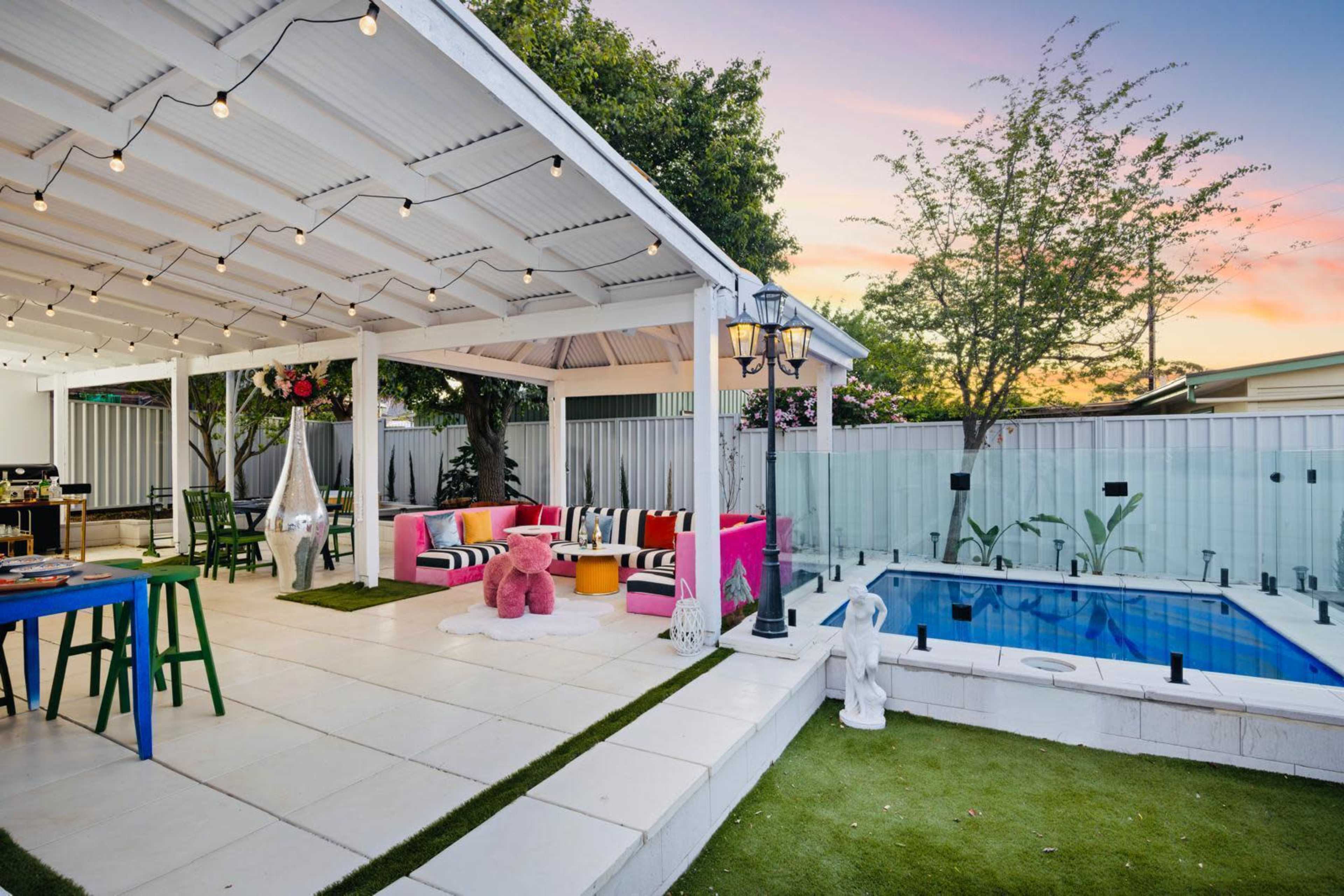 The image shows an outdoor patio area with colorful seating, a small pool, and string lights overhead, surrounded by greenery.