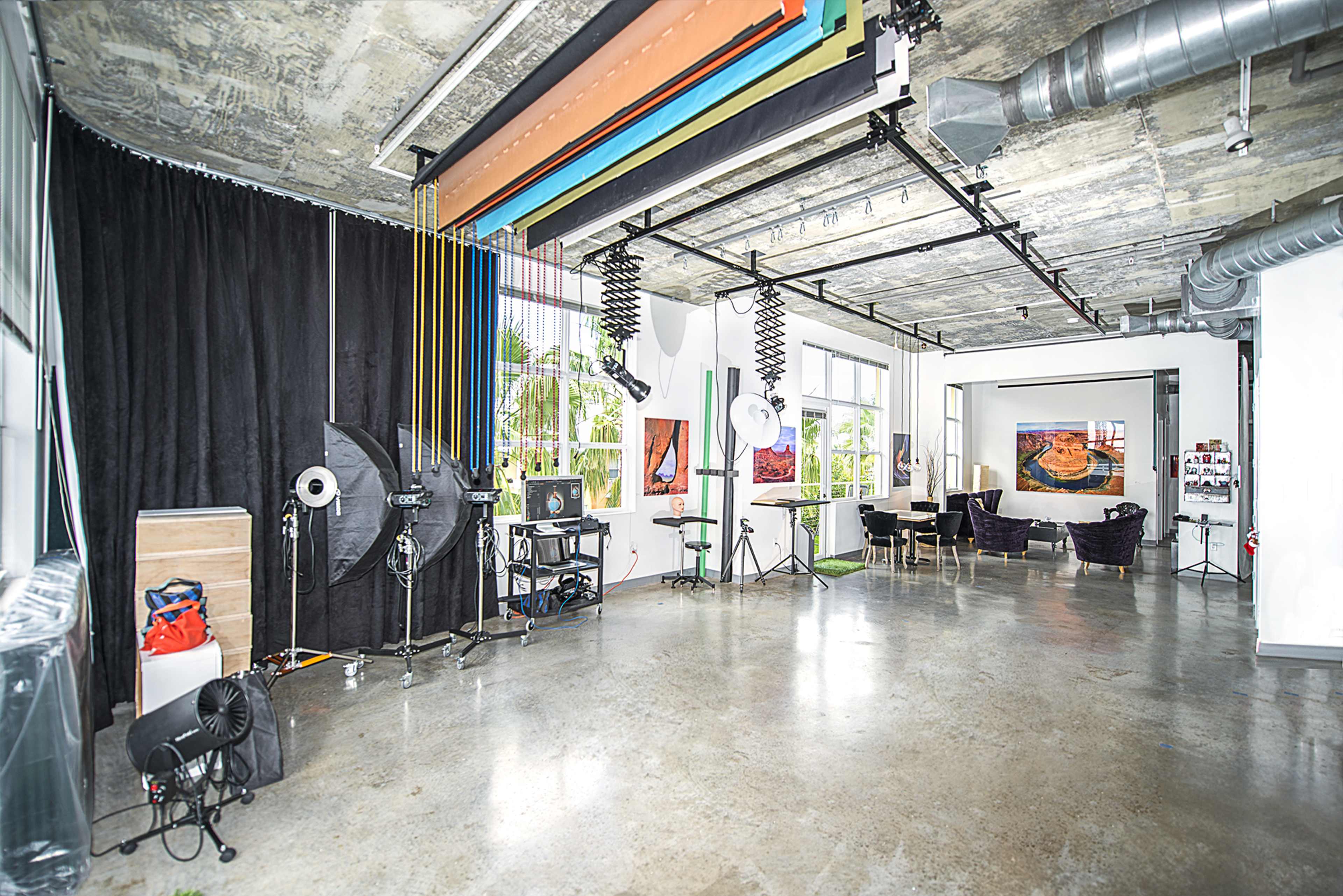 The image shows a spacious, modern studio with various lighting equipment, a seating area, and colorful backdrops hanging from the ceiling.