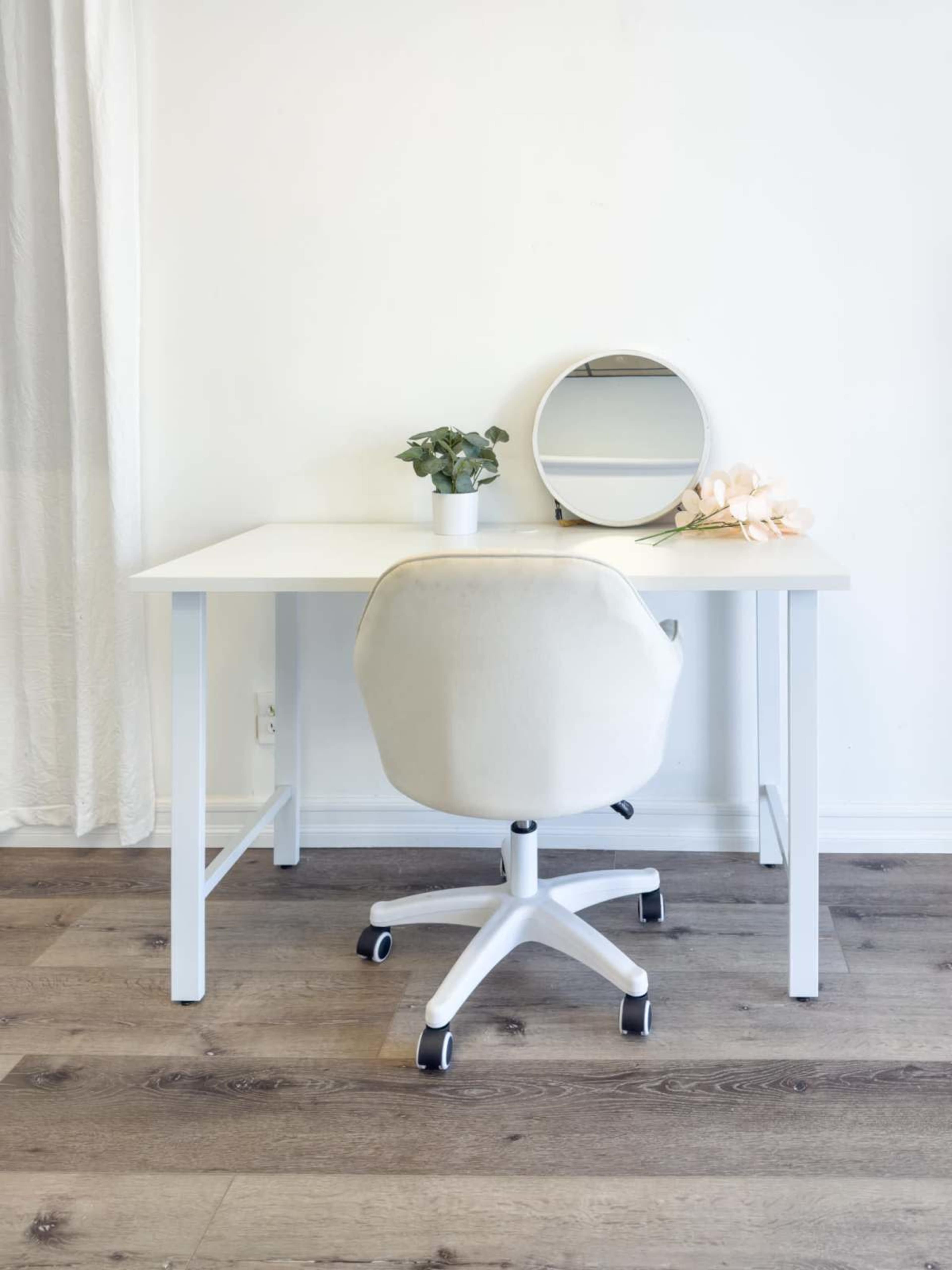A white desk with a round mirror, a small potted plant, and a light-colored office chair is set against a plain wall.