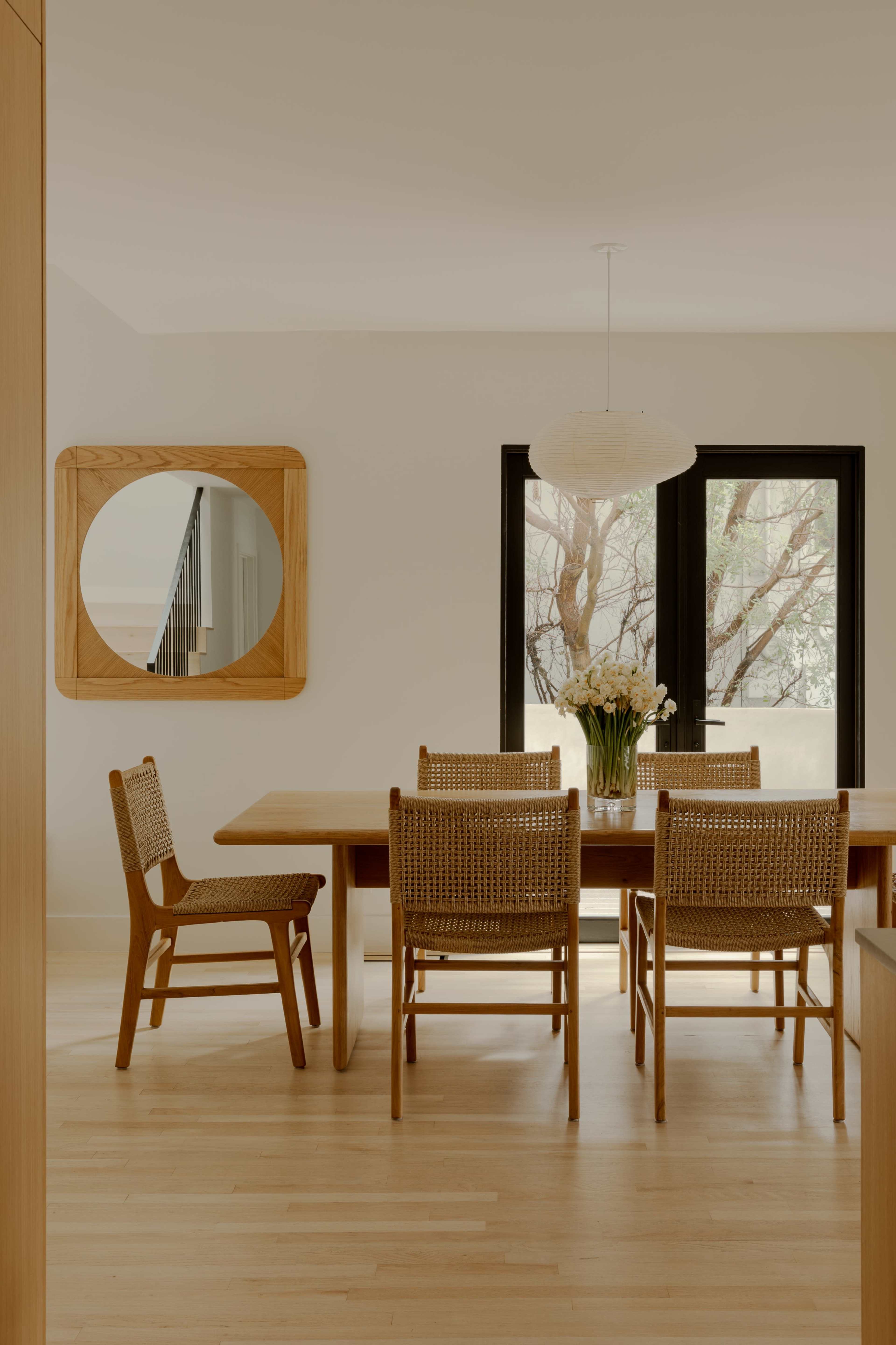 A modern dining room features a wooden table surrounded by chairs, with a large mirror and a window showcasing tree branches outside.