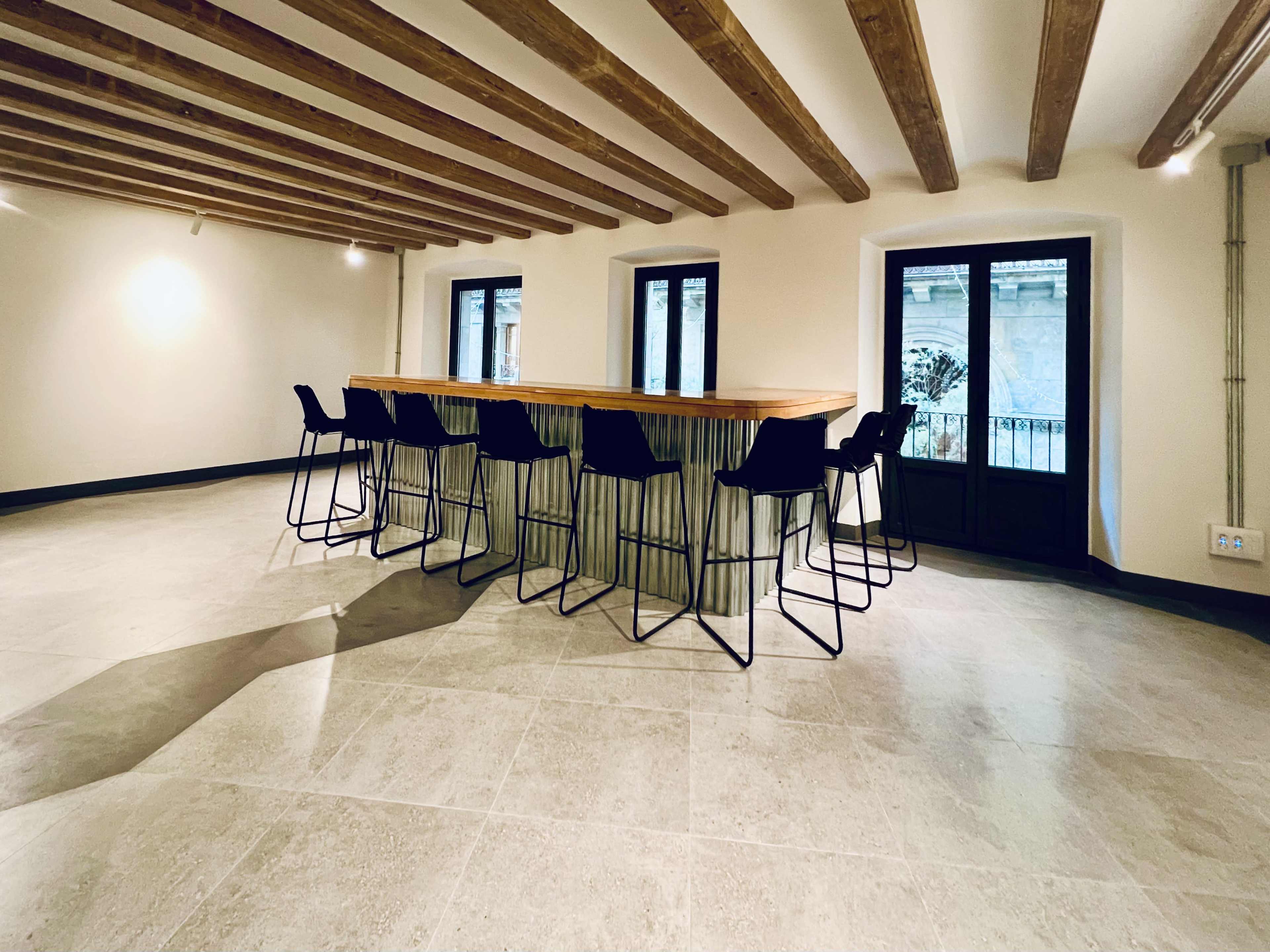 The image shows a spacious room with a long wooden bar and high stools, featuring large windows and exposed wooden beams on the ceiling.