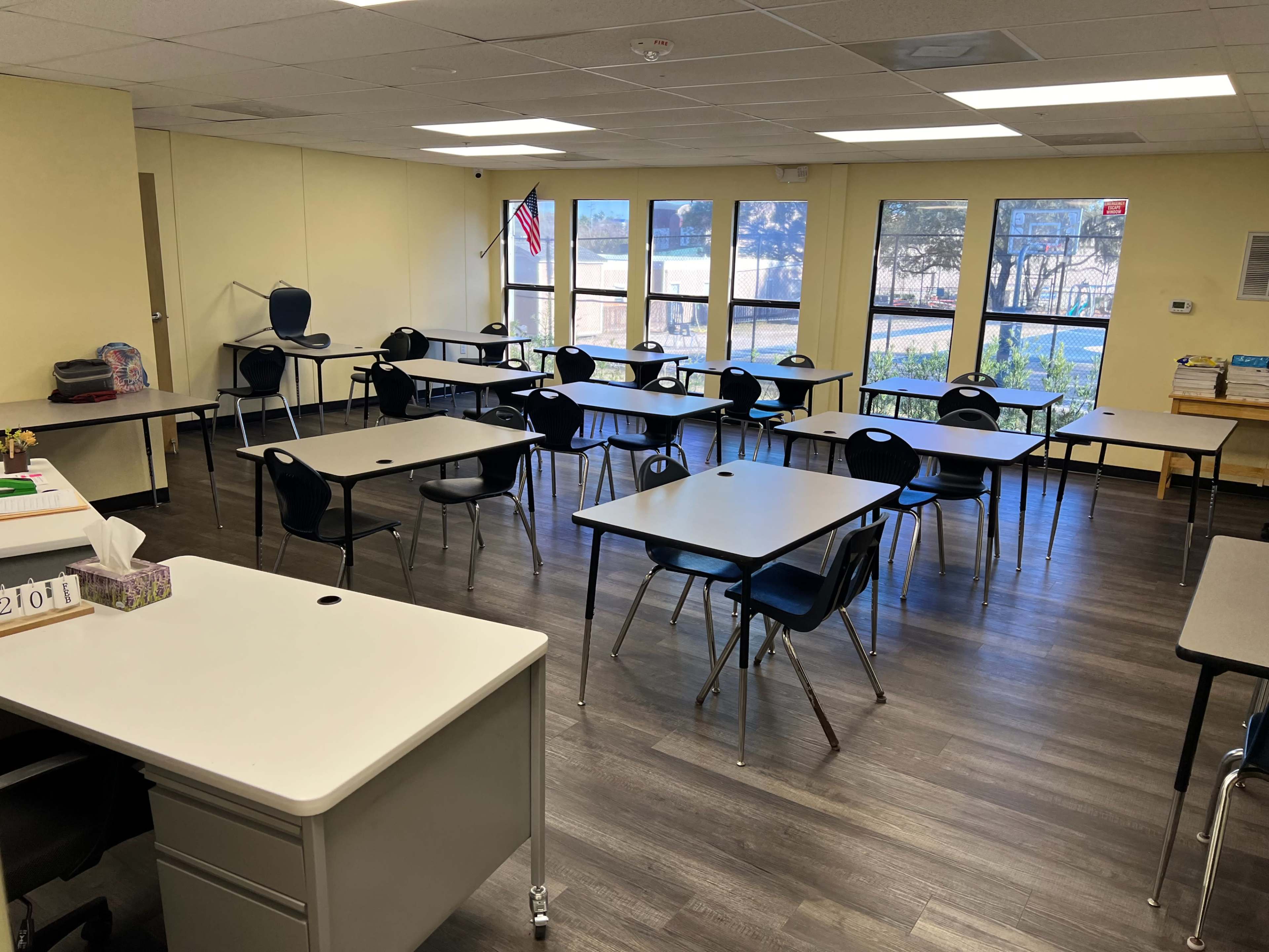 A classroom is arranged with multiple tables and chairs, featuring windows that overlook an outdoor area, and a desk positioned in the foreground.