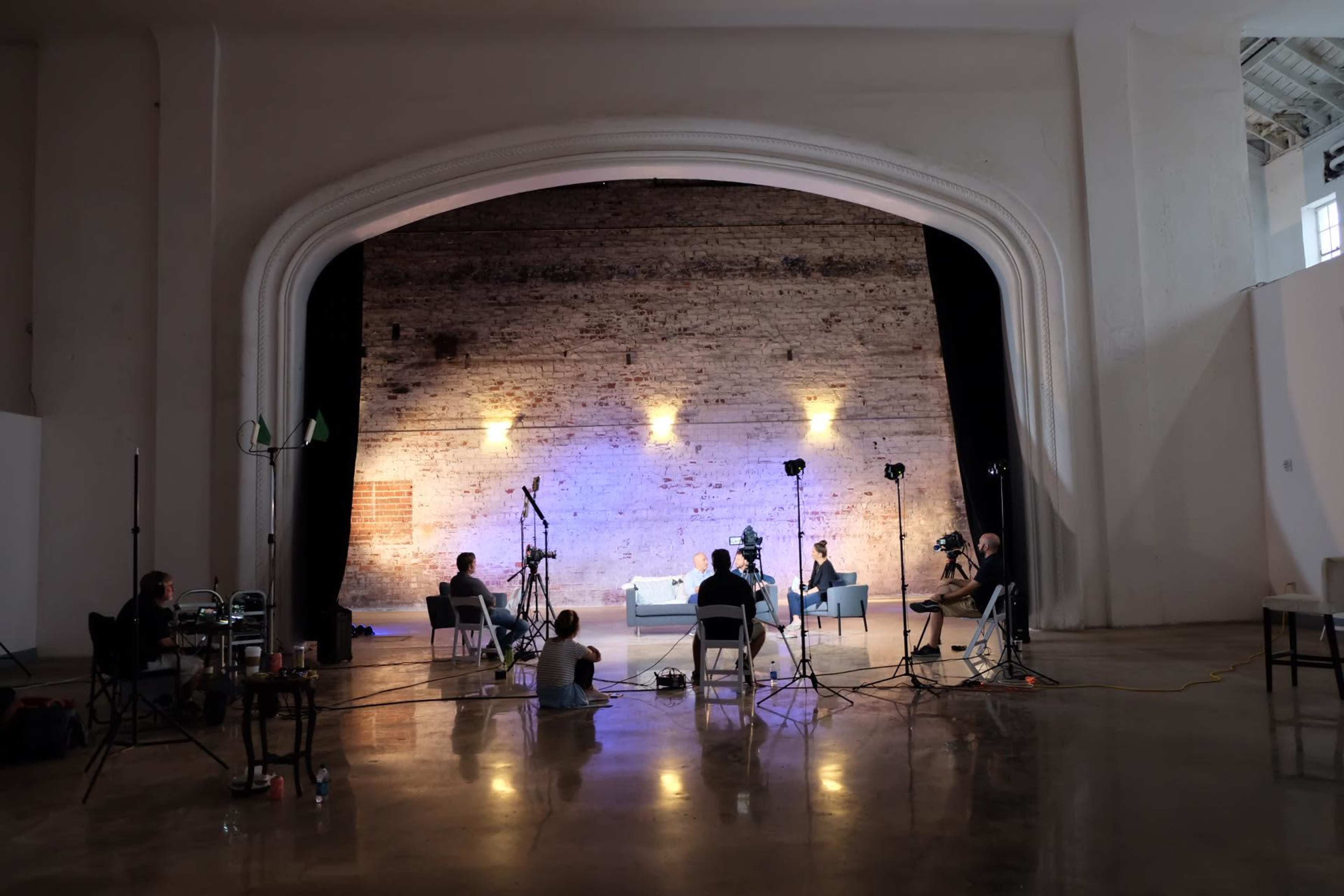 A film crew is set up in a spacious, well-lit room with a large, arched backdrop and several cameras pointed at a seated interviewee.