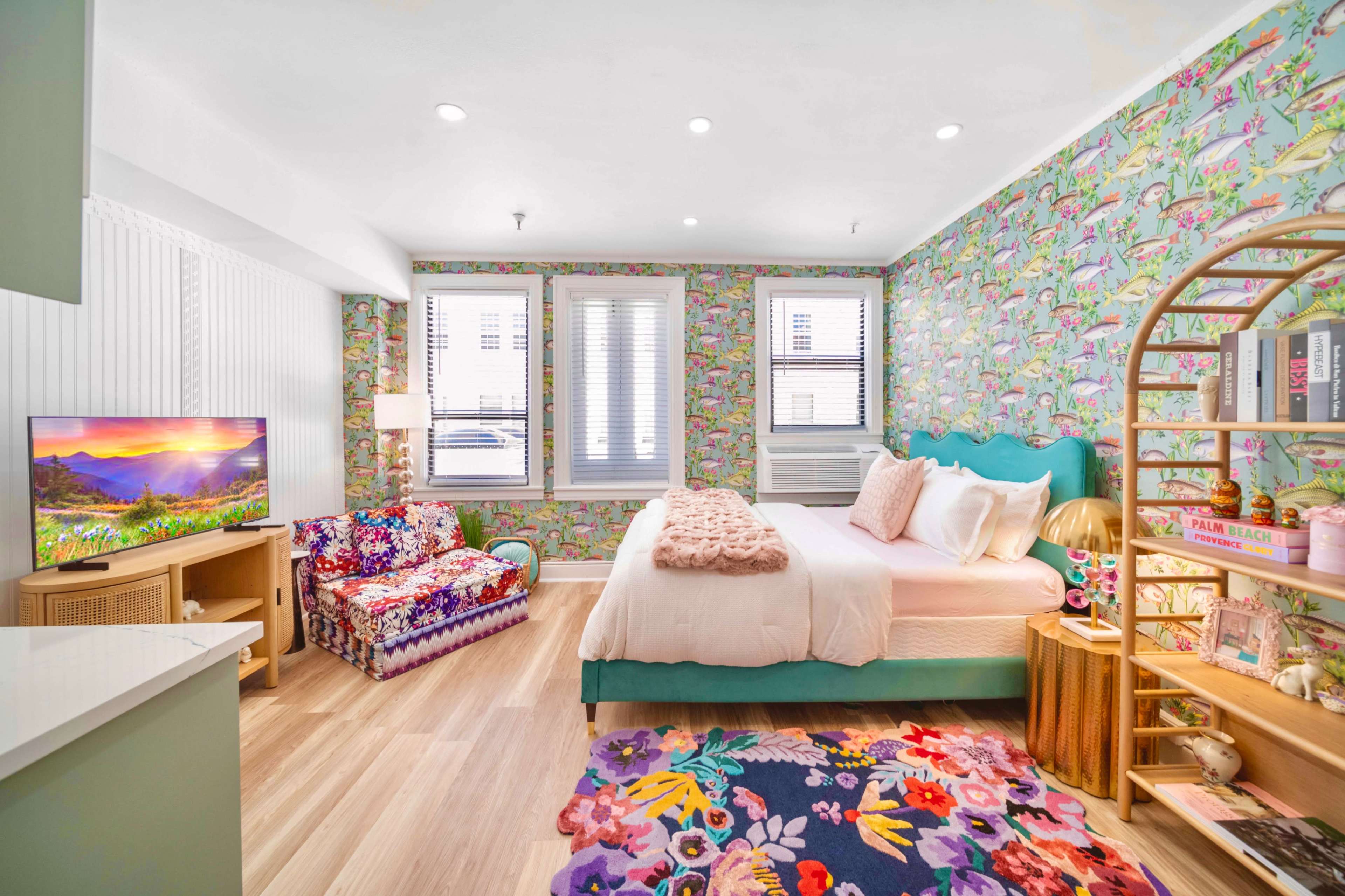 A bright bedroom with floral wallpaper, a bed with a teal headboard, a colorful seating area, and a television.