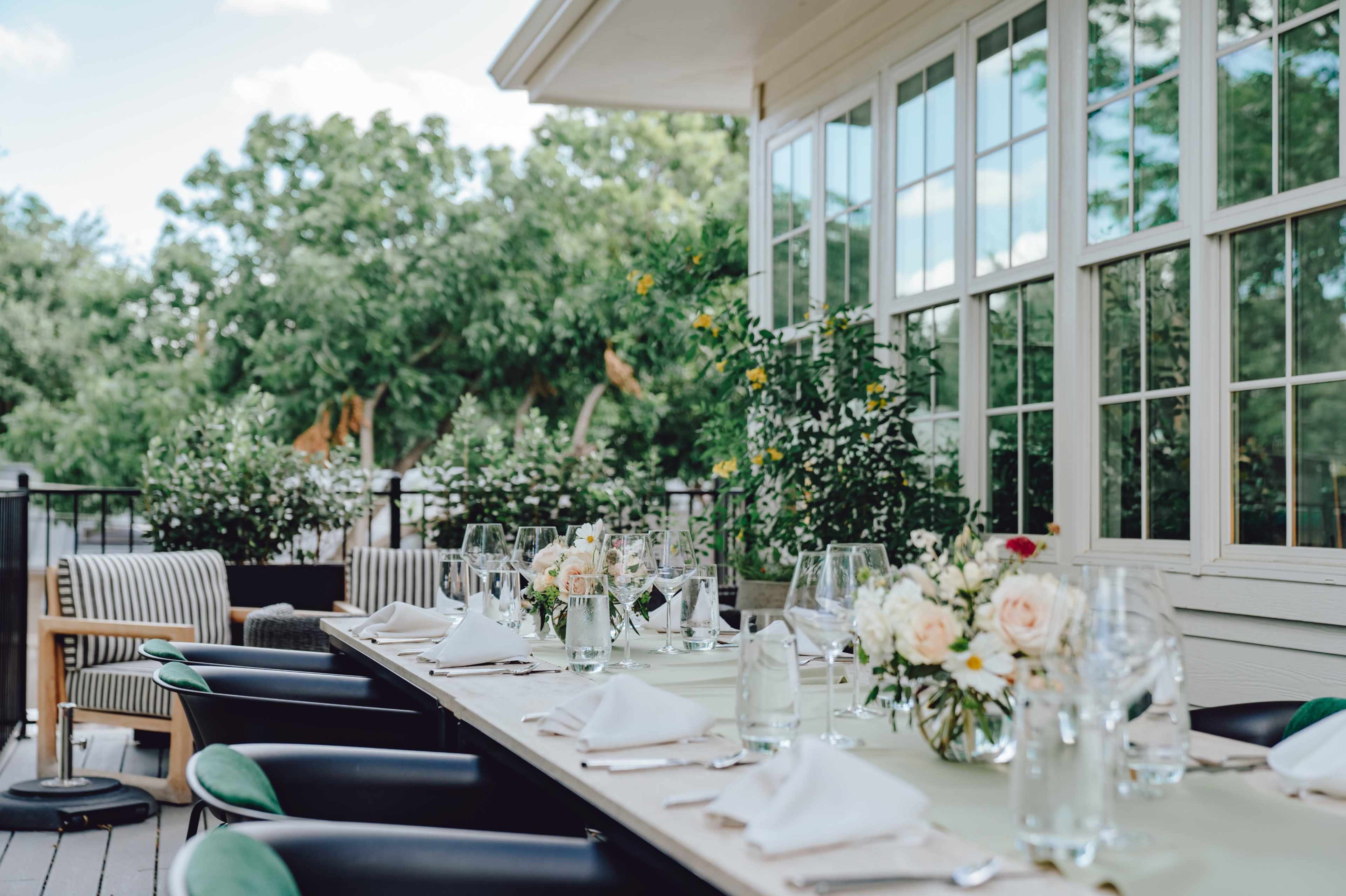 A beautifully arranged dining table is set outdoors, featuring dishes, glassware, and floral centerpieces, with greenery visible in the background.