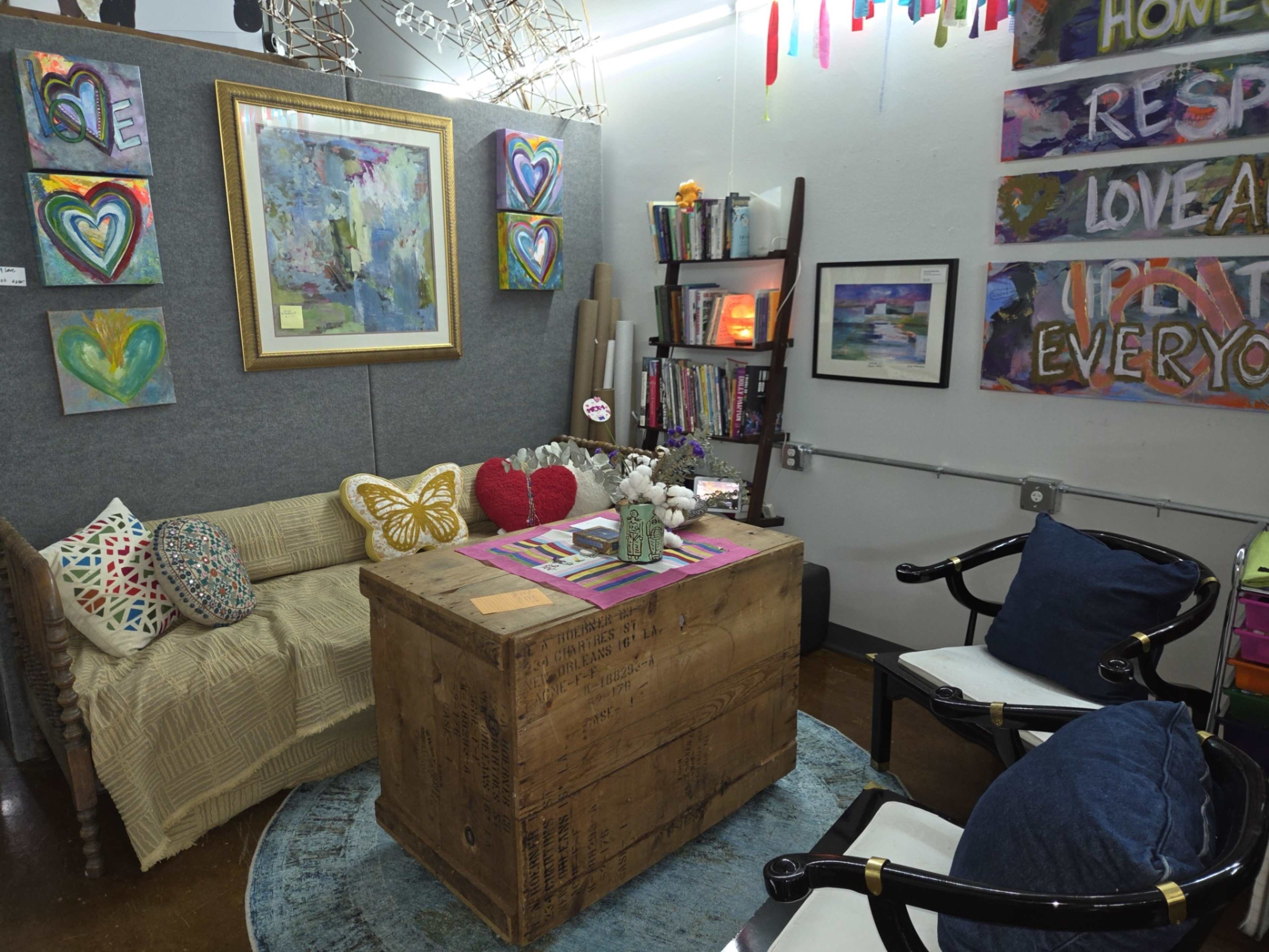 The image displays a cozy art studio featuring a wooden table at the center, surrounded by a couch and chairs, with colorful paintings and decor on the walls.