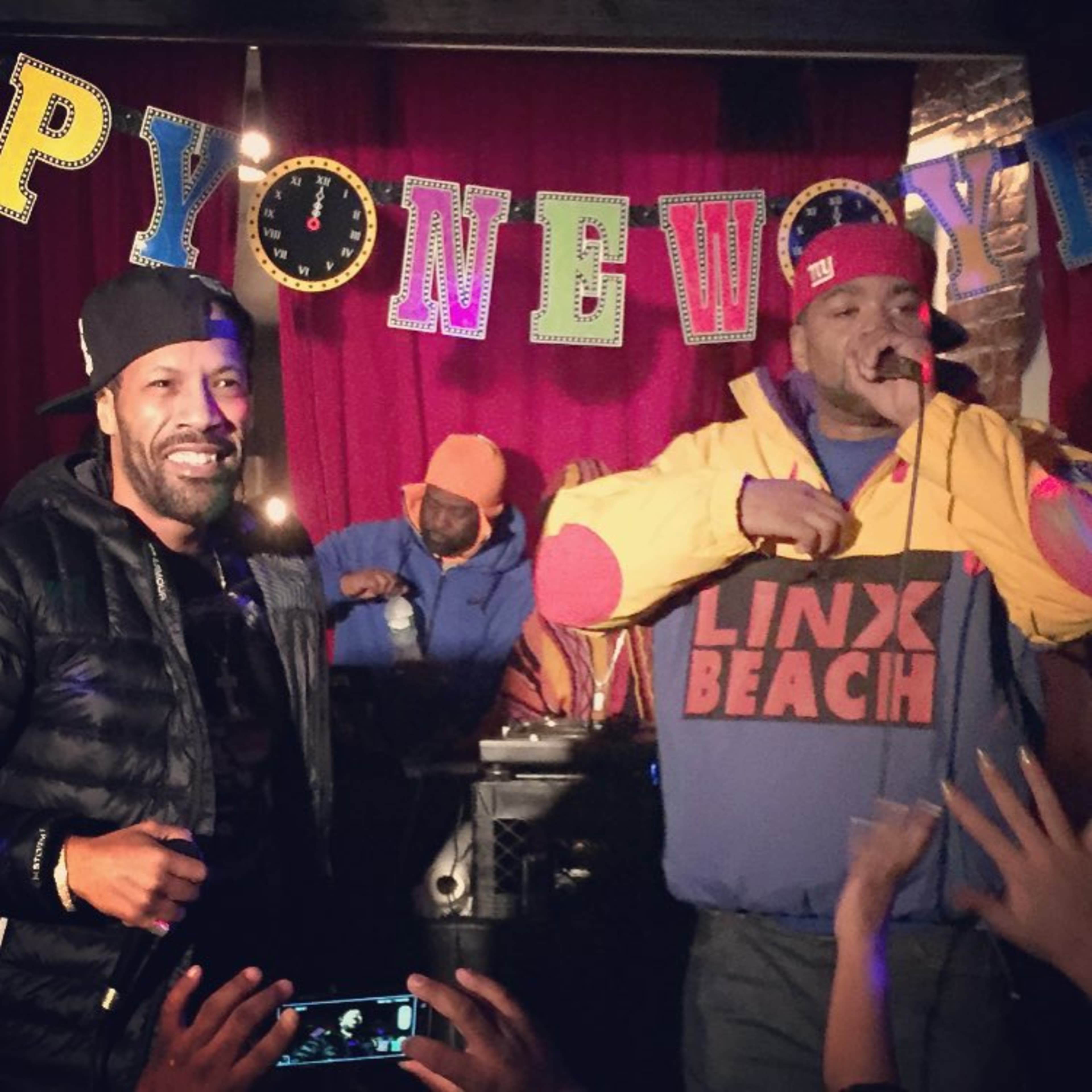 Two male hip-hop artists perform on stage with a festive banner in the background while a crowd engages with them.