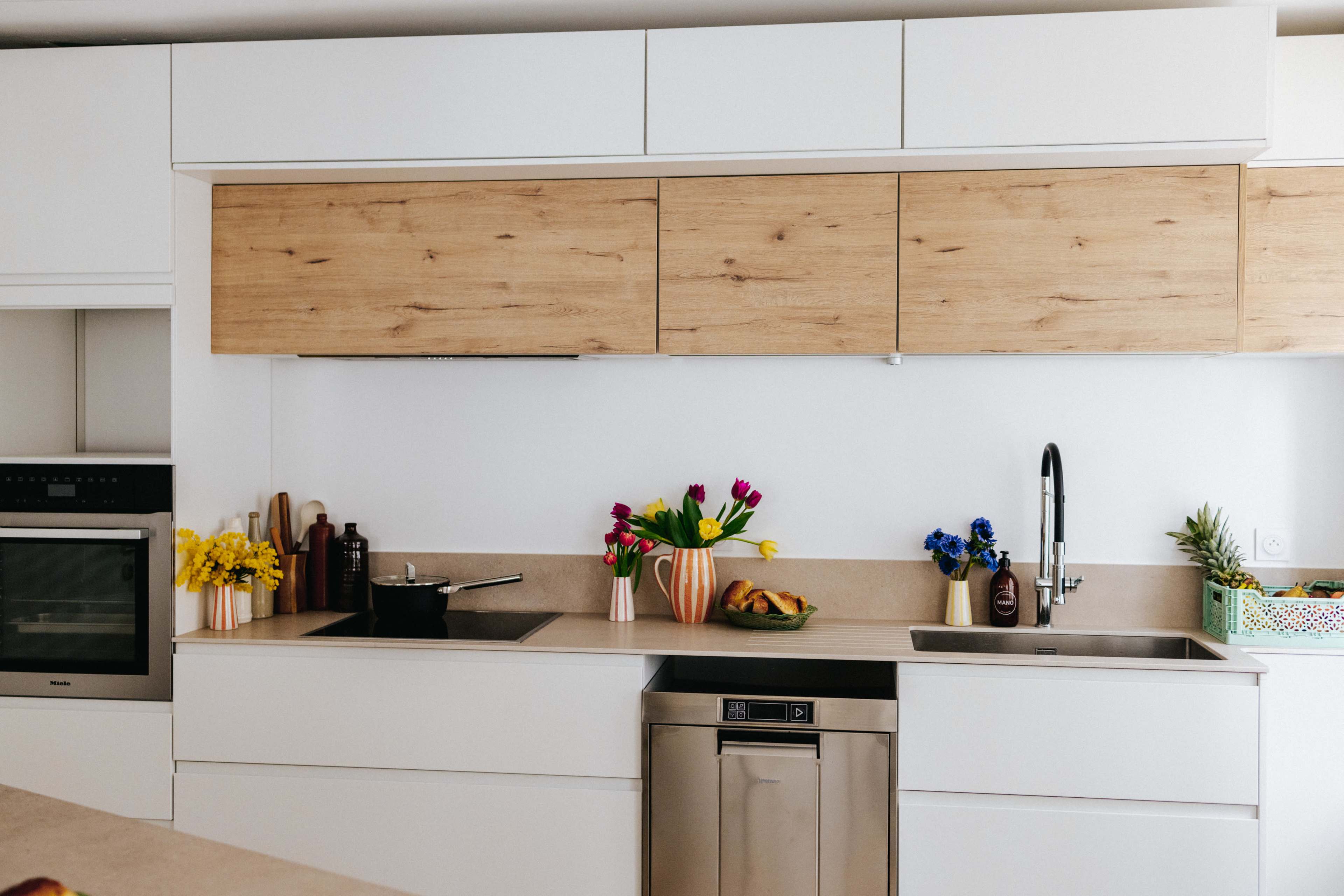 A modern kitchen features wooden cabinets, a stainless steel dishwasher, a sink, and vases of fresh flowers on the countertop.