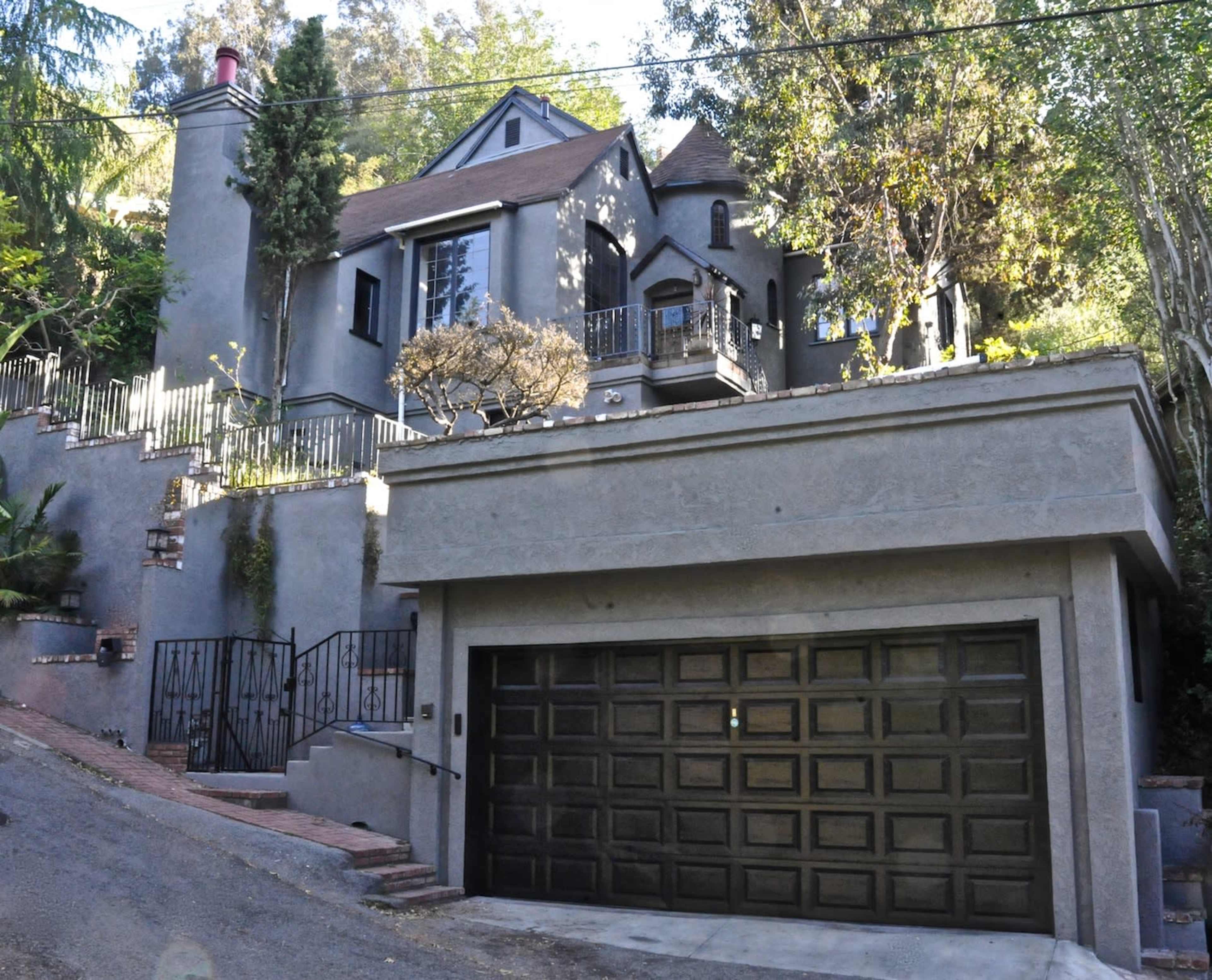 A gray two-story house with a balcony and a garage is situated on a hillside, surrounded by trees.