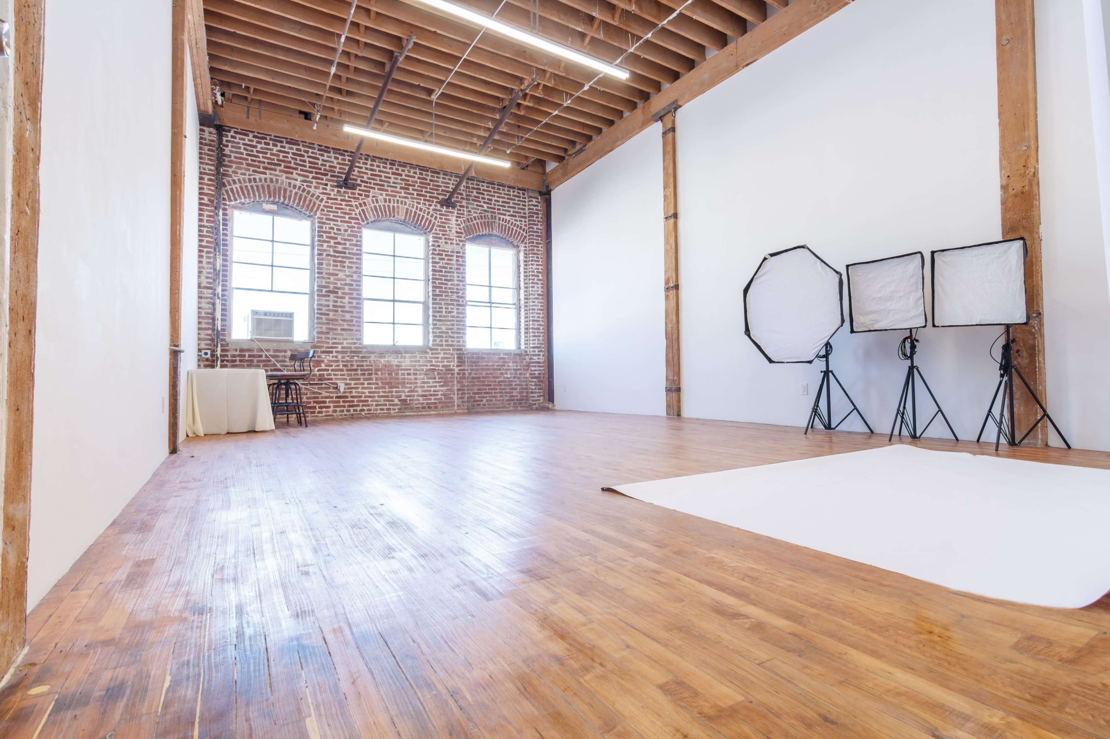 A spacious photography studio with wooden floors, exposed brick walls, and lighting equipment in the corner.