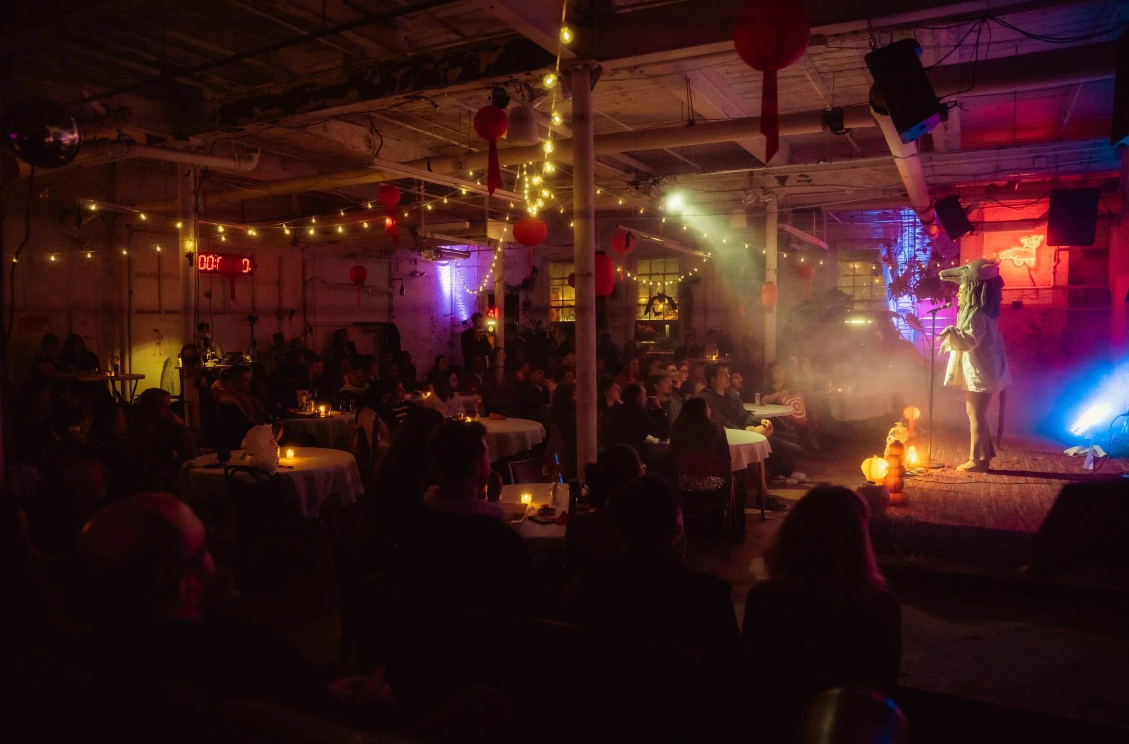 A performer stands on stage in a dimly lit venue filled with tables of seated audience members, surrounded by colorful lights and decorations.