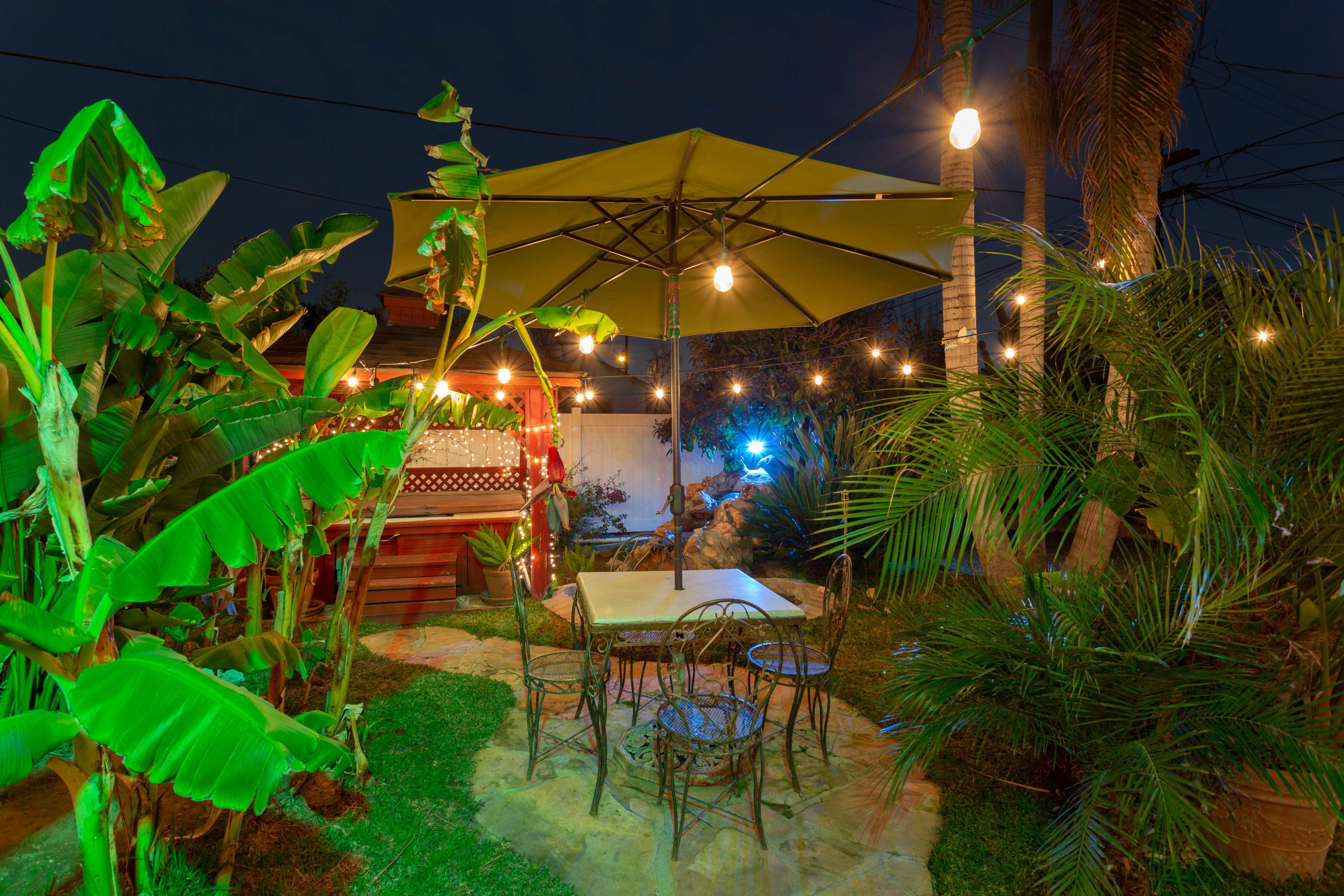 A patio area features a large yellow umbrella over a table and chairs, surrounded by tropical plants and illuminated by string lights.