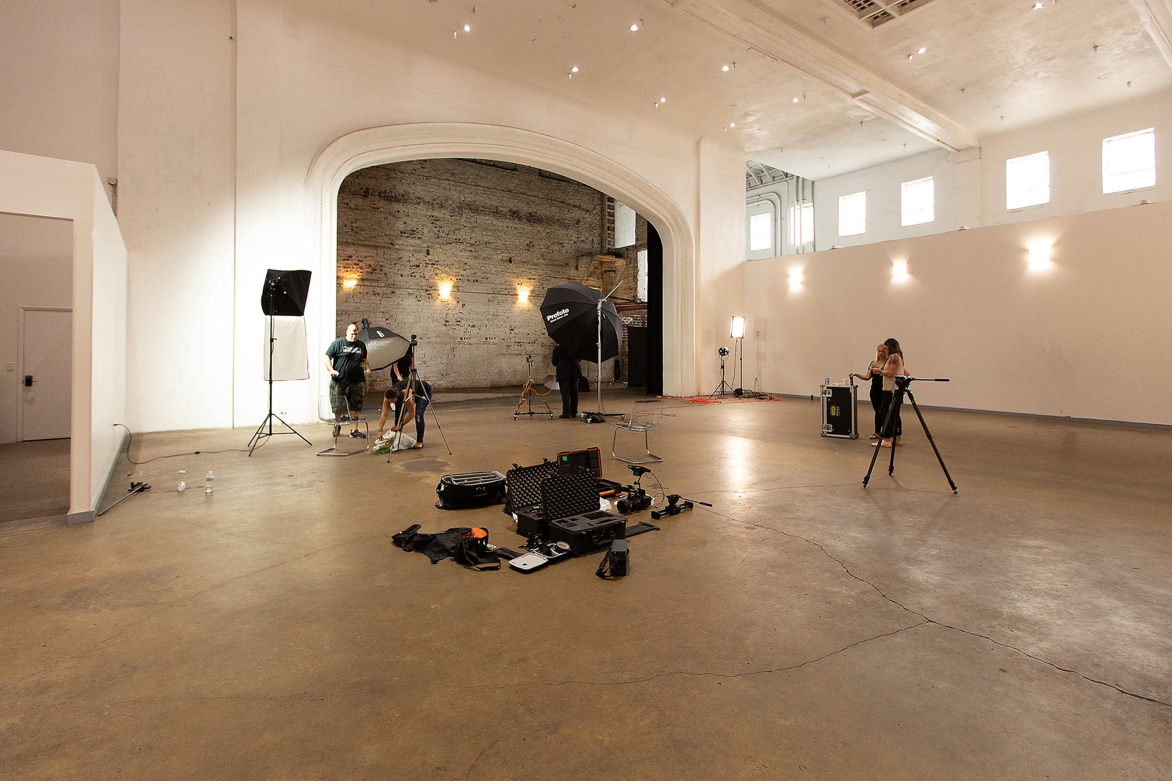 A large, empty studio space is set up for a photoshoot, featuring lighting equipment and a photographer preparing their gear.