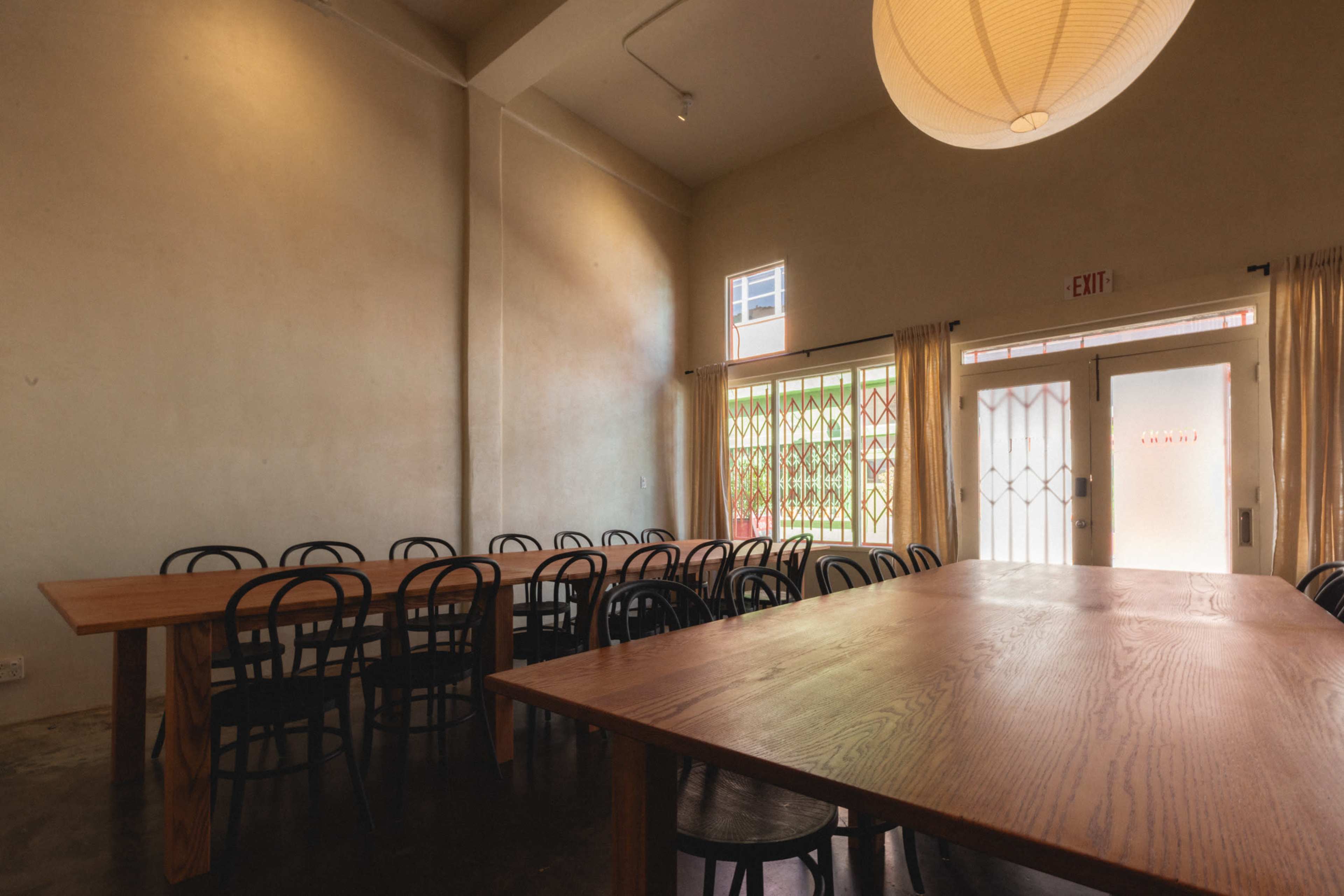 A spacious, well-lit room features long wooden tables arranged with black chairs, complemented by large windows and minimalist decor.