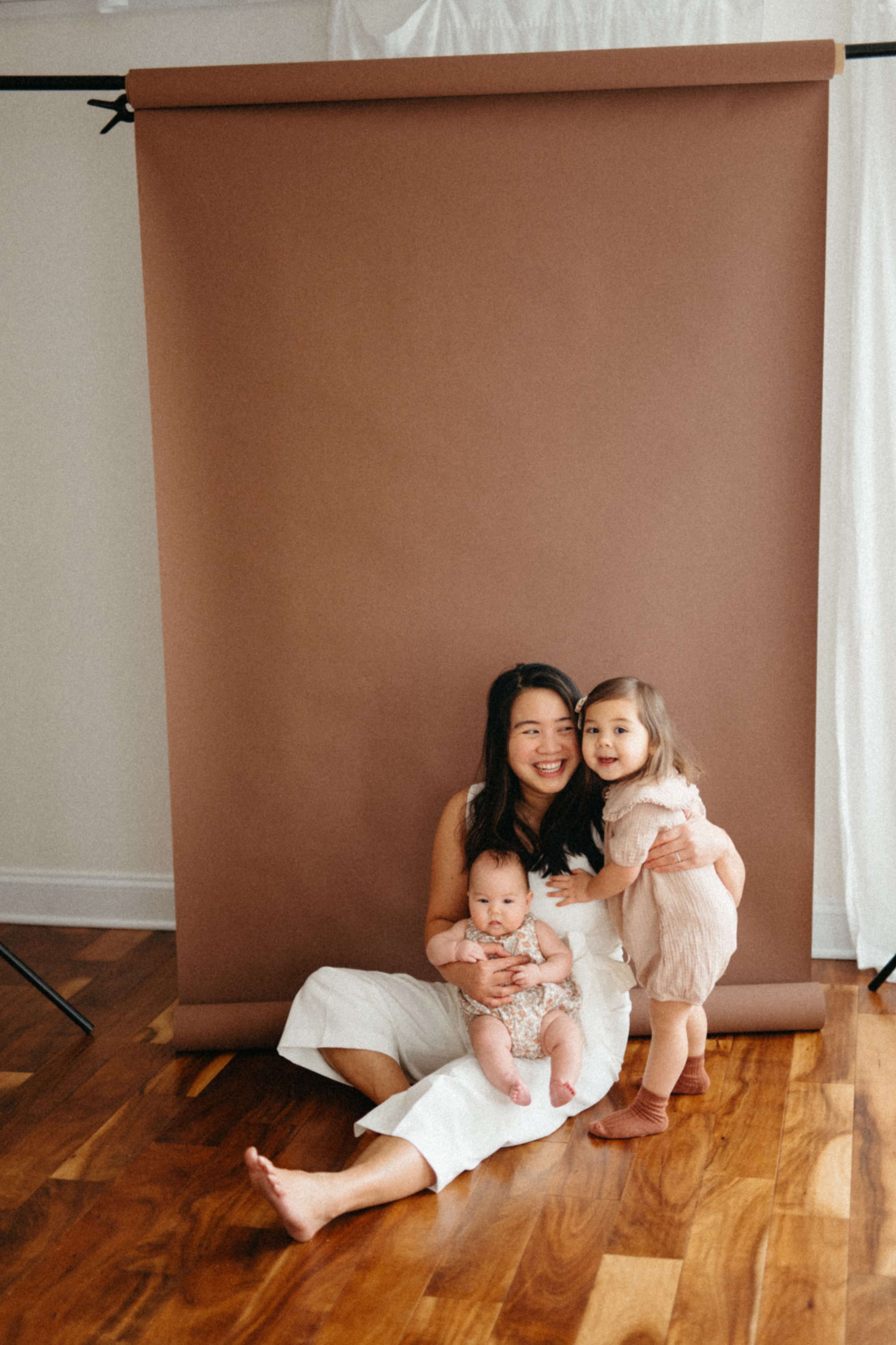 A woman sits on the floor with a baby in her lap and a young girl standing beside her, against a brown backdrop.