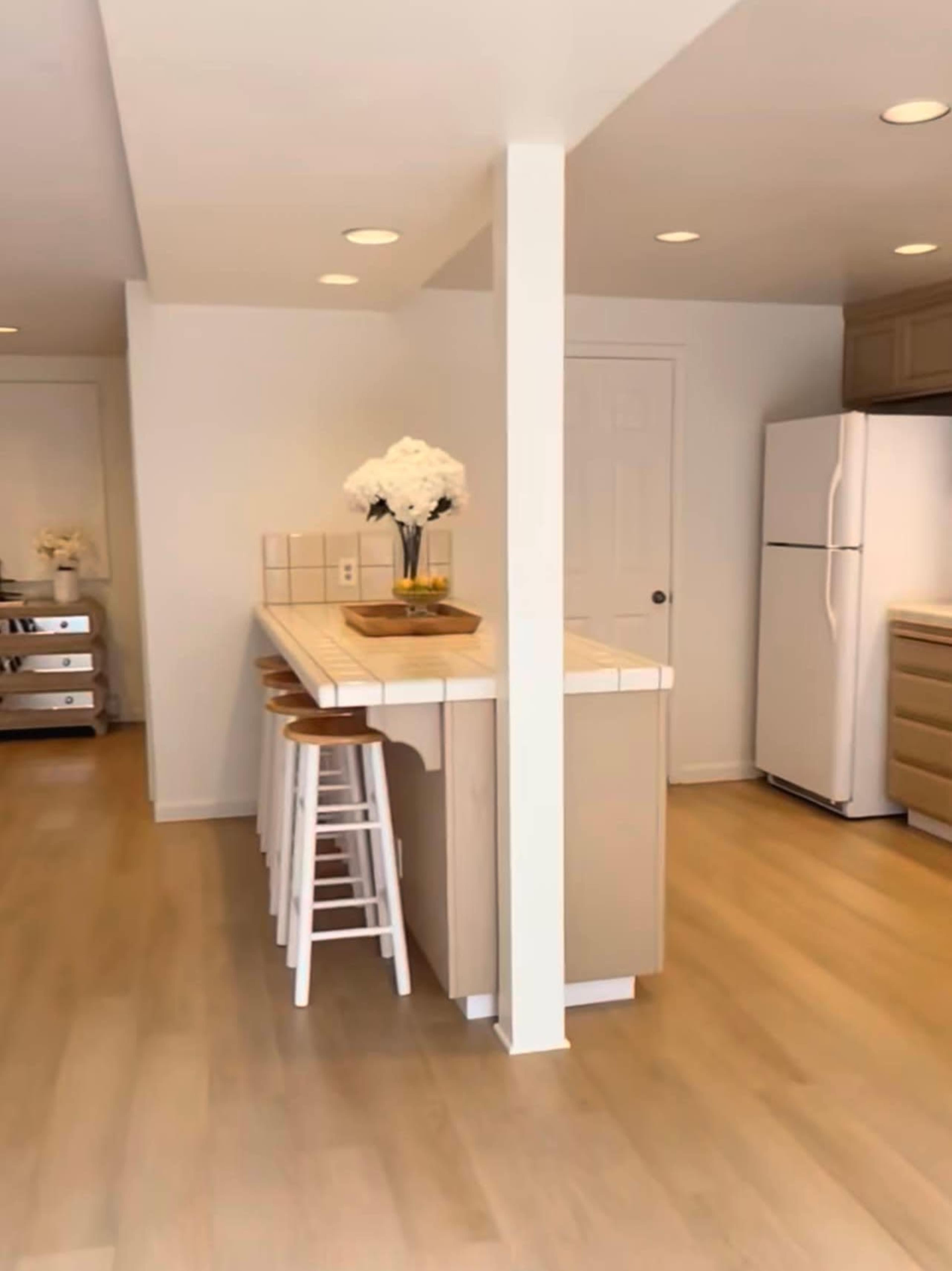 The image shows a modern kitchen with a tiled island, bar stools, and white cabinetry, featuring a refrigerator and natural light.