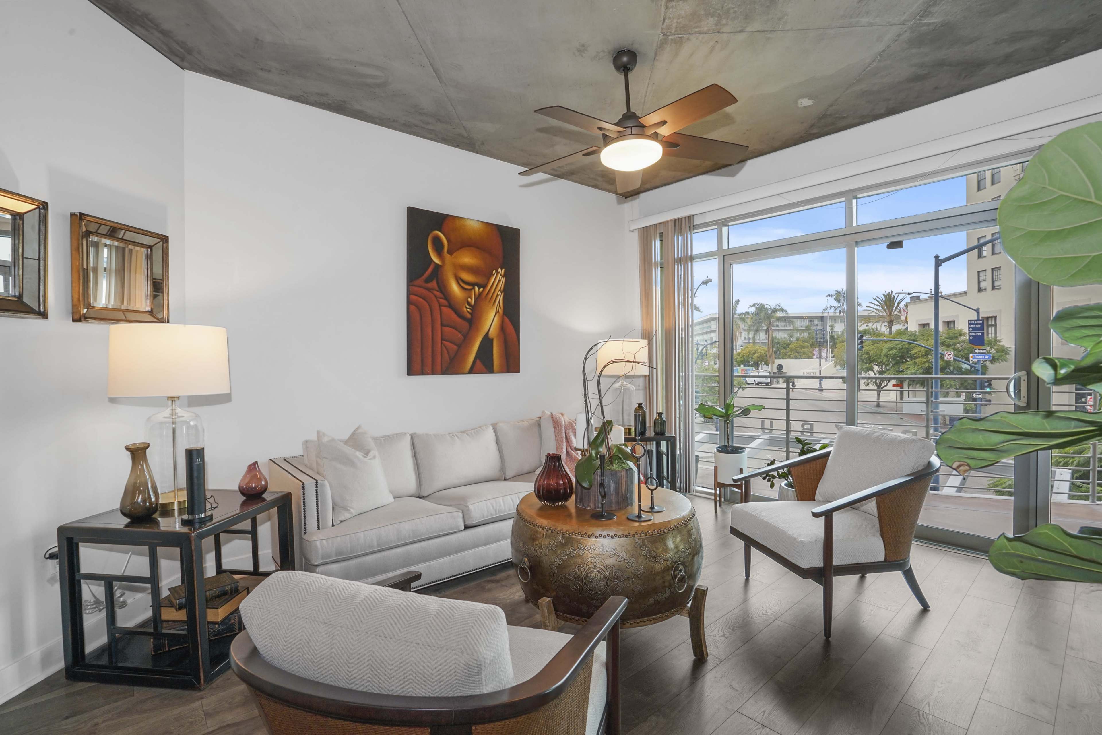 A modern living room features a light-colored sofa, wooden chairs, a round coffee table, and large windows with views of an outdoor setting.