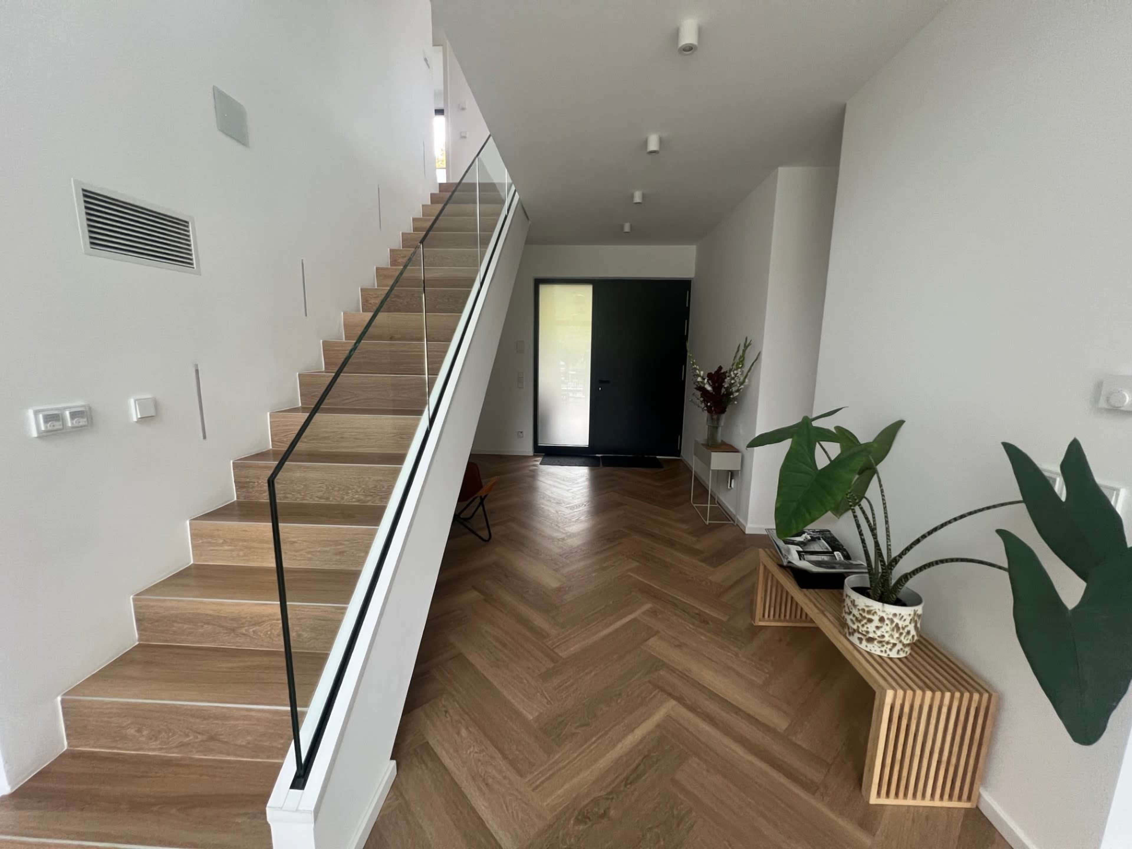 The image shows a modern interior hallway featuring a staircase with glass railings, a dark front door, and herringbone-patterned wooden flooring.
