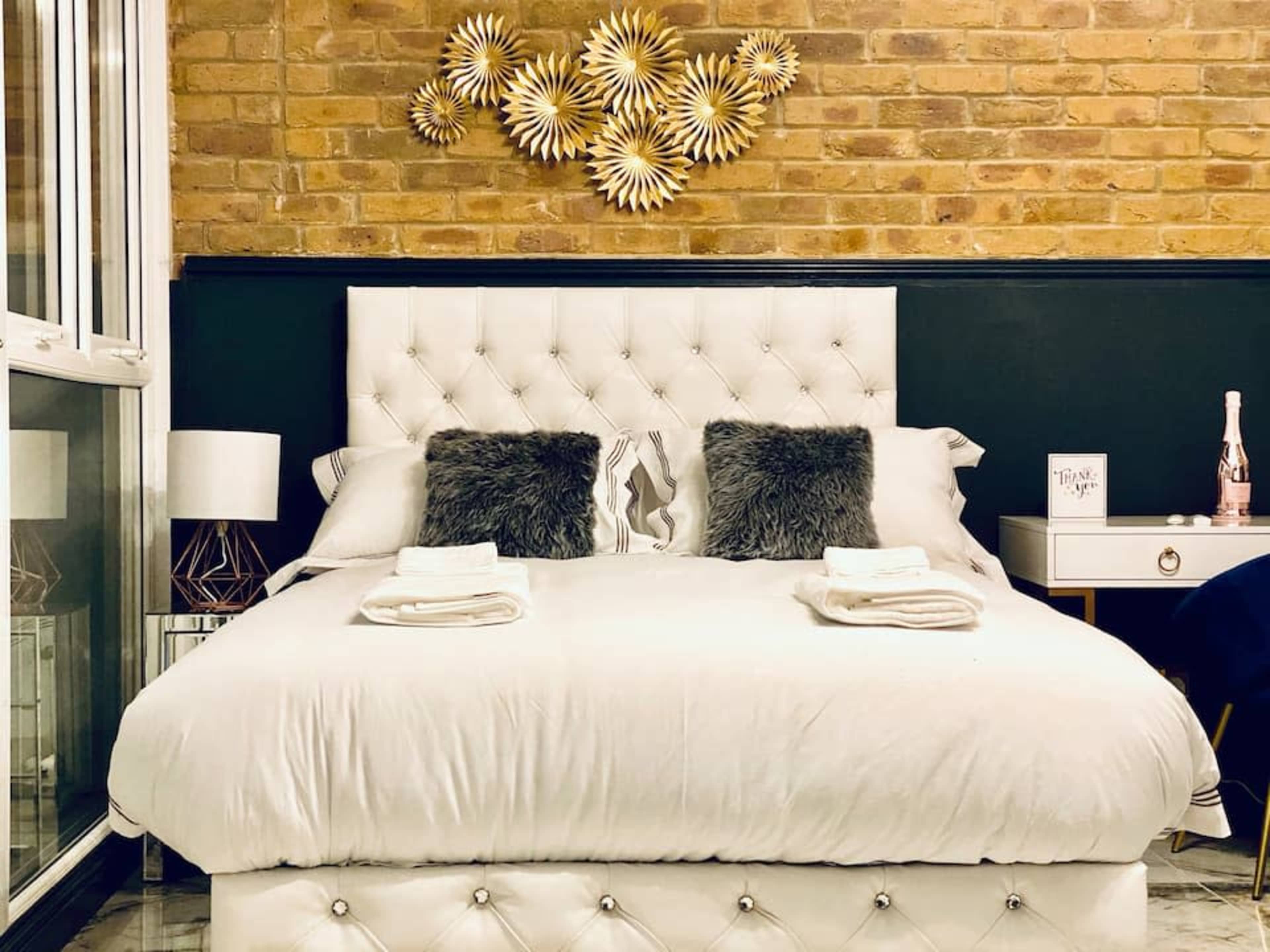 A neatly arranged bedroom features a tufted white bed with two decorative pillows, a bedside table, and a wall adorned with a sunburst art piece.