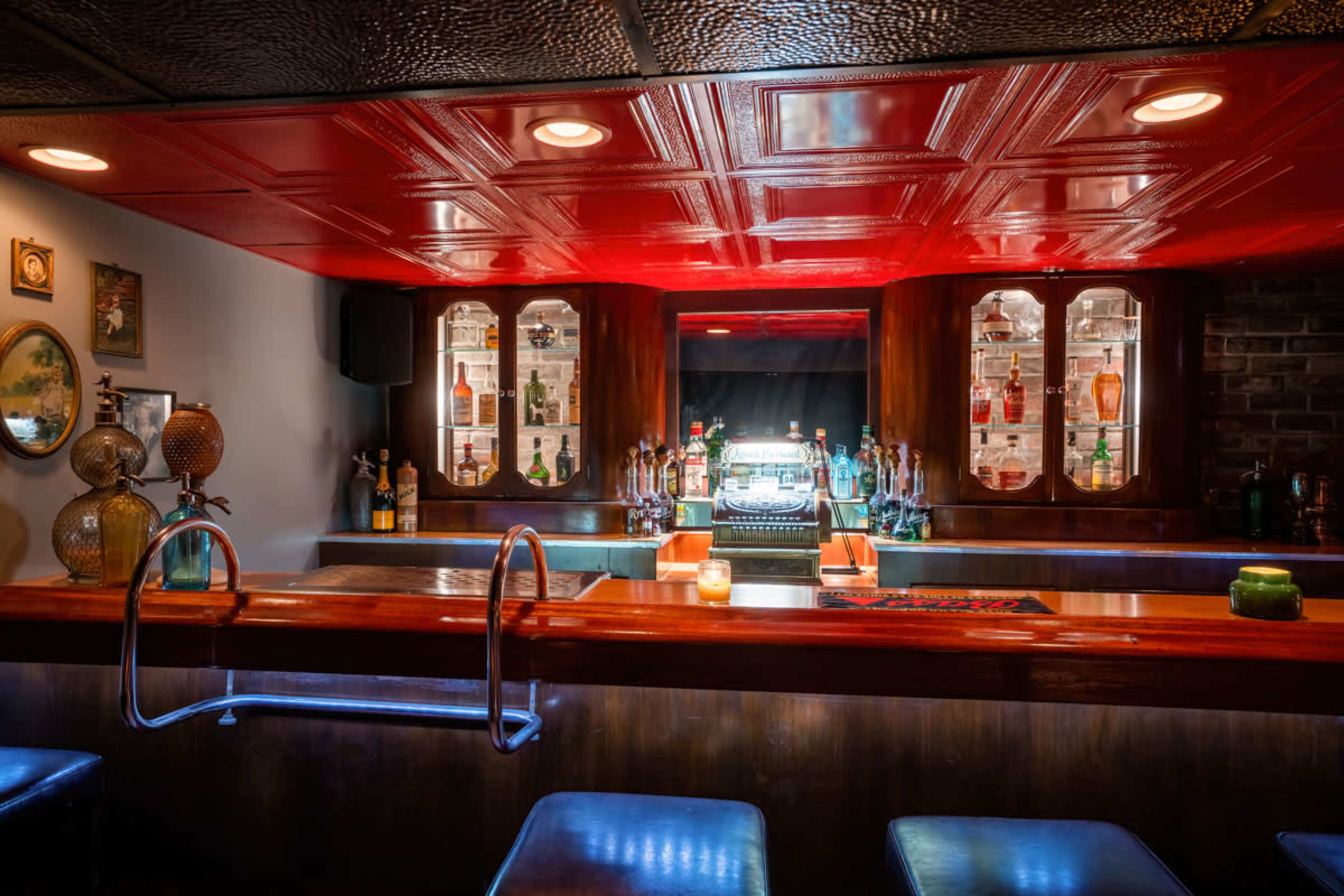 A well-lit bar with a wooden counter, illuminated bottles on display, and a red ceiling.