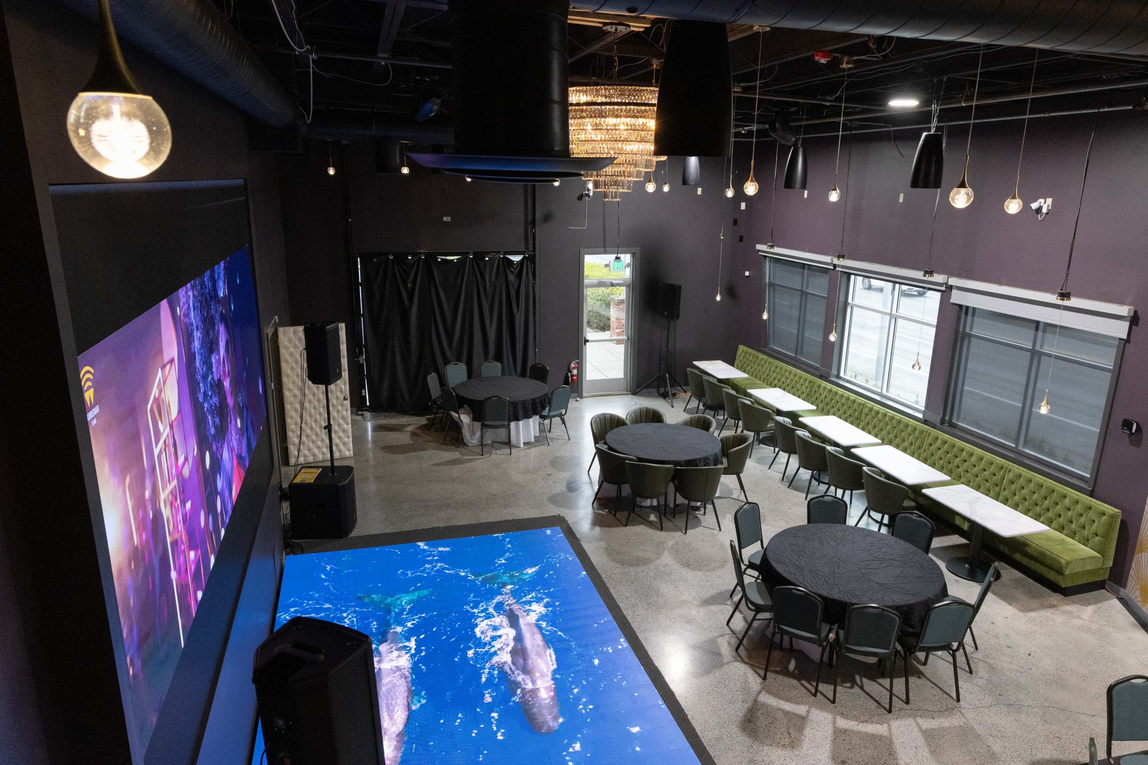 The image shows a modern event space featuring round and rectangular tables, a large projection screen displaying aquatic visuals, and a long green sofa along one wall.