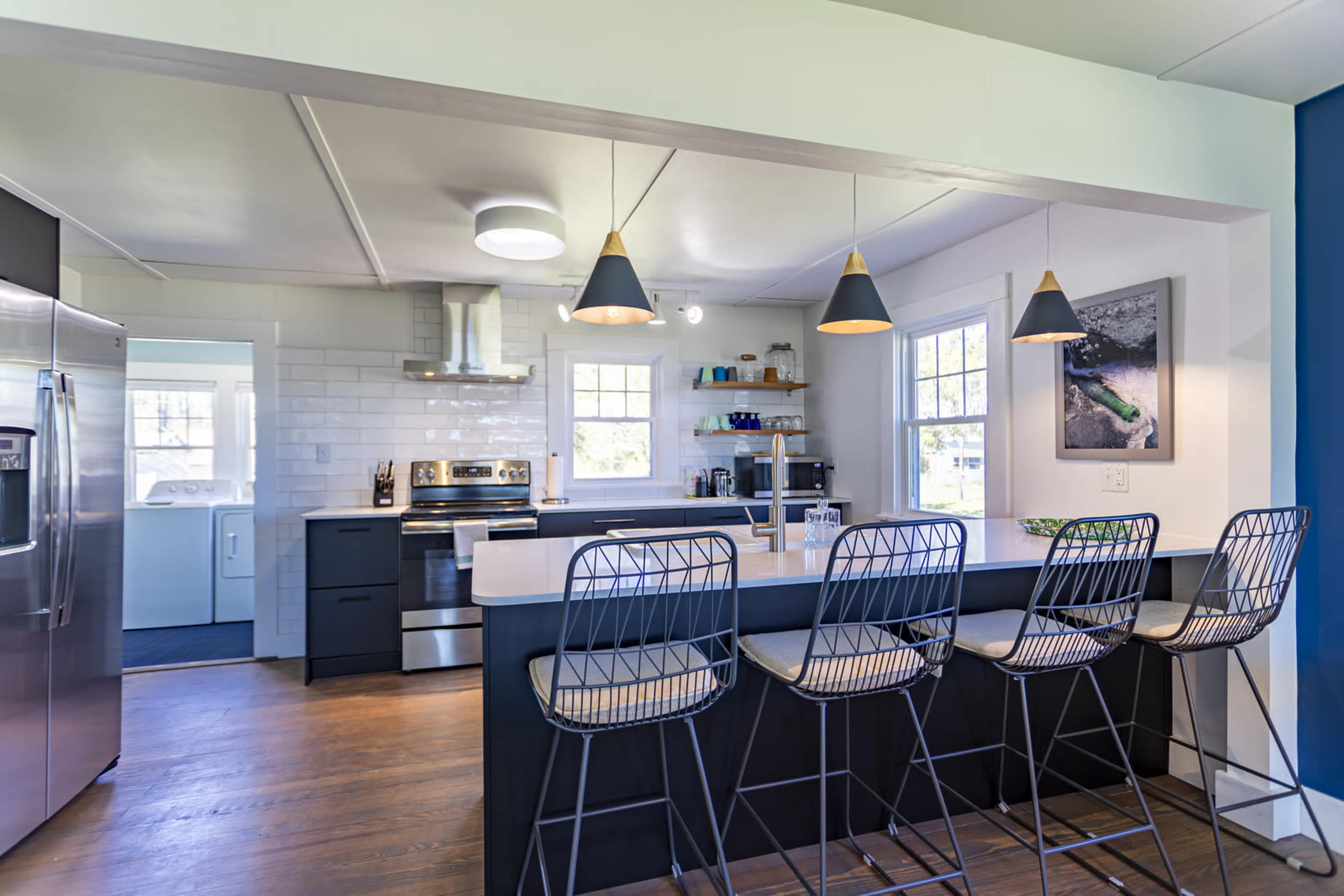 The image shows a modern kitchen with stainless steel appliances, white cabinetry, and four wire bar stools positioned at a dark counter.