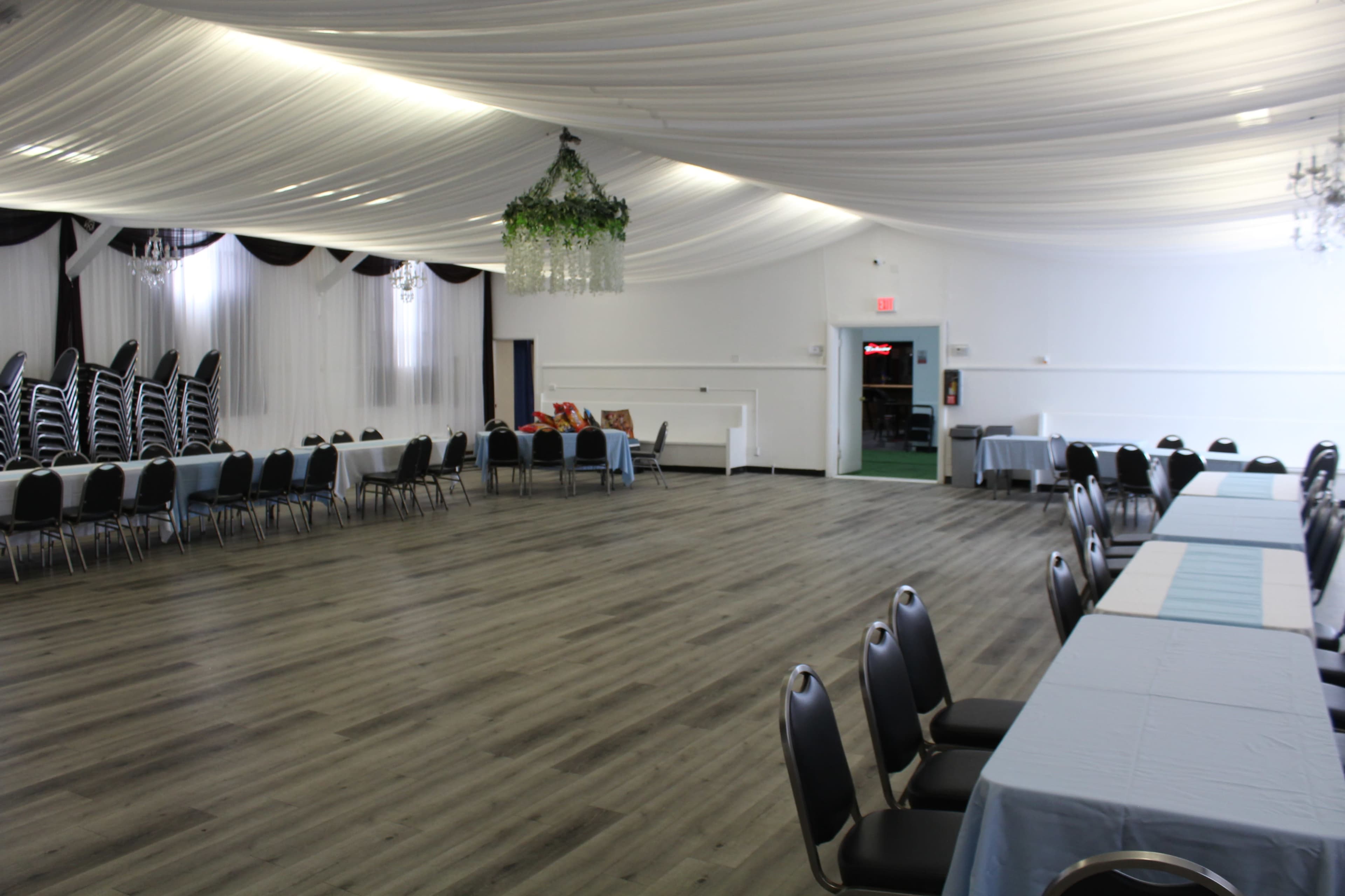 The spacious event hall features a draped ceiling with chandeliers, arranged seating along the sides, and a row of stacked chairs at the back.