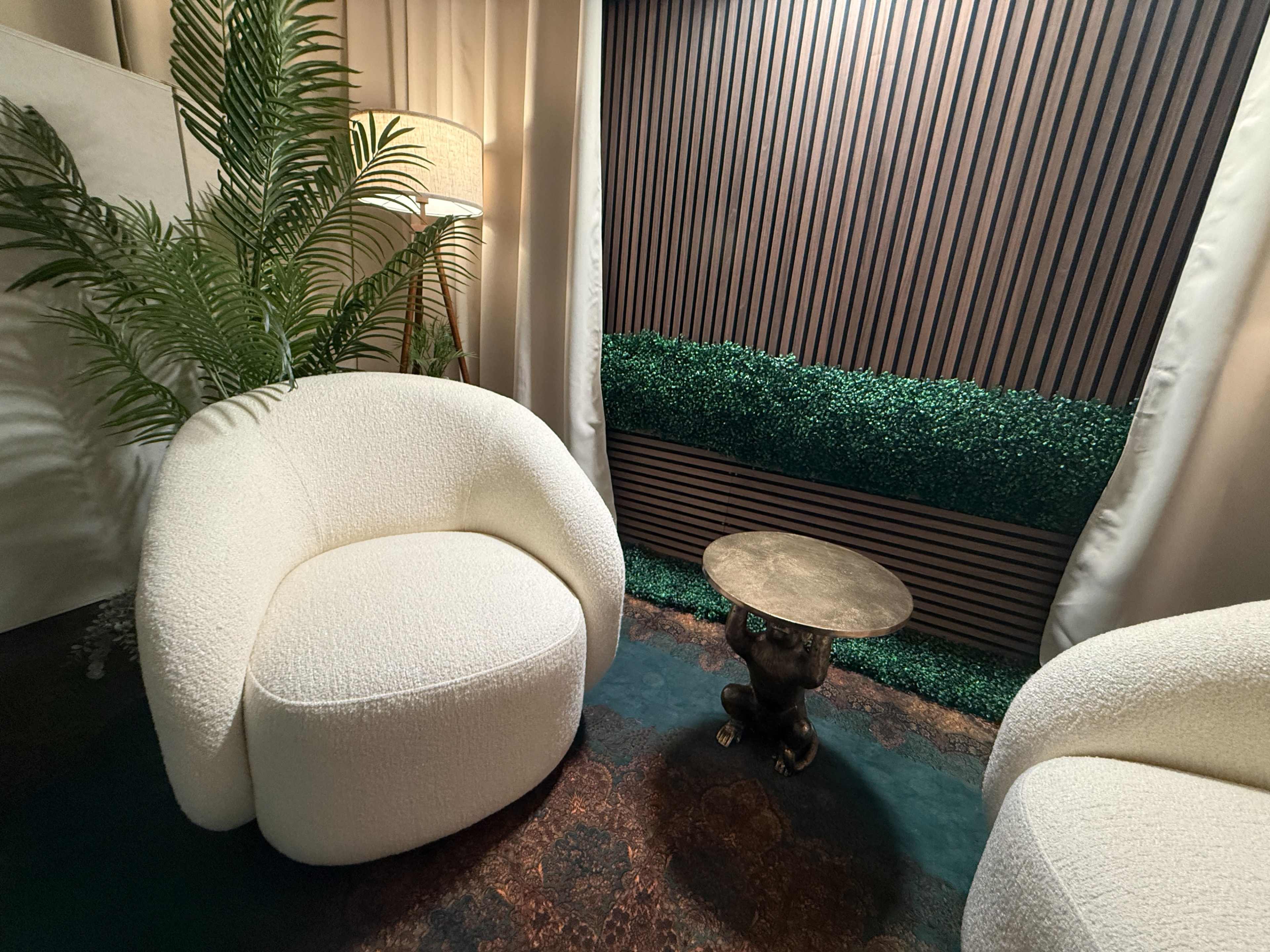 The scene features two round, white chairs beside a small brass table, with a backdrop of green foliage and textured wooden panels.