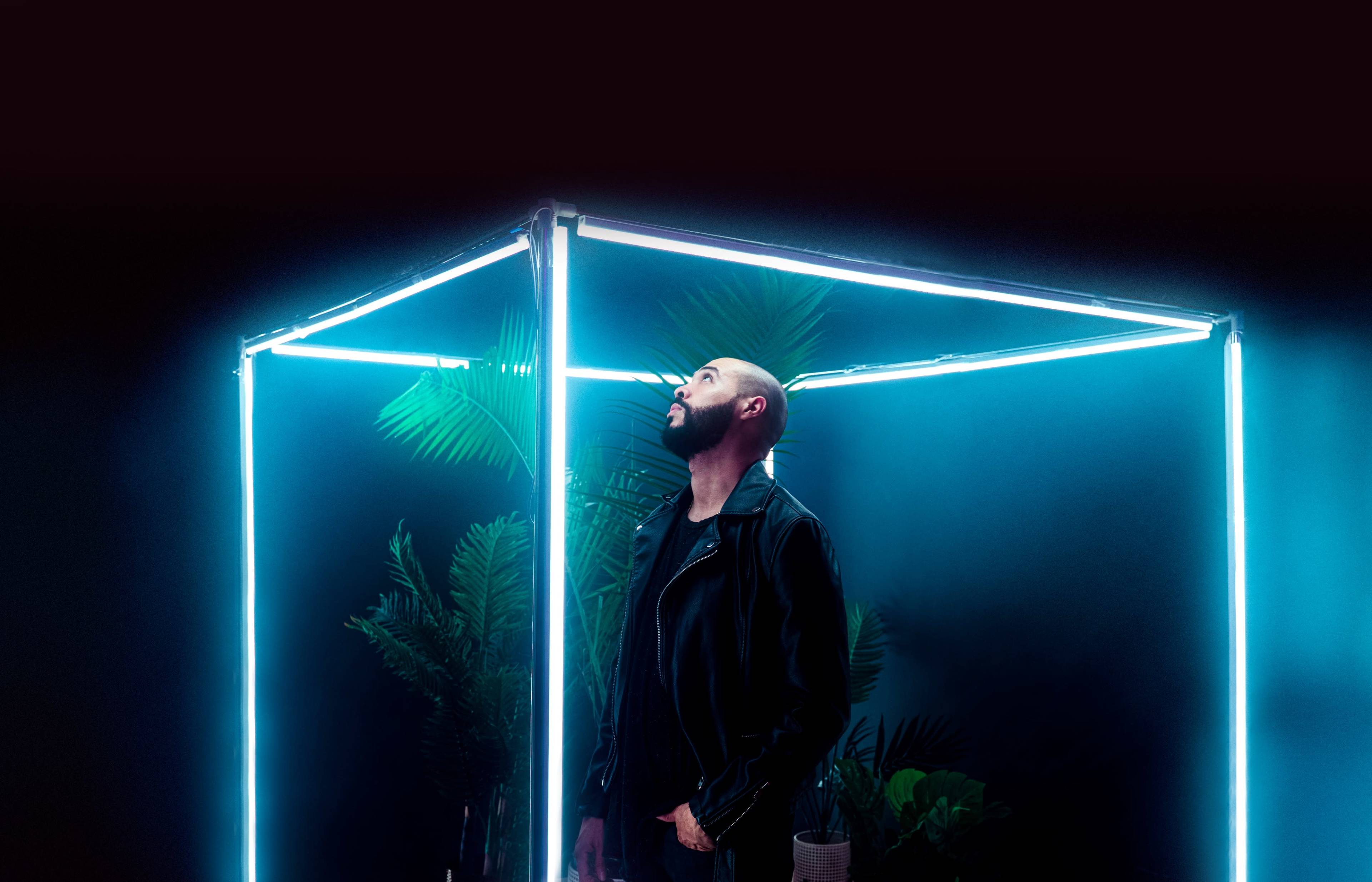 A man stands inside a geometric structure illuminated by blue neon lights, surrounded by plants.