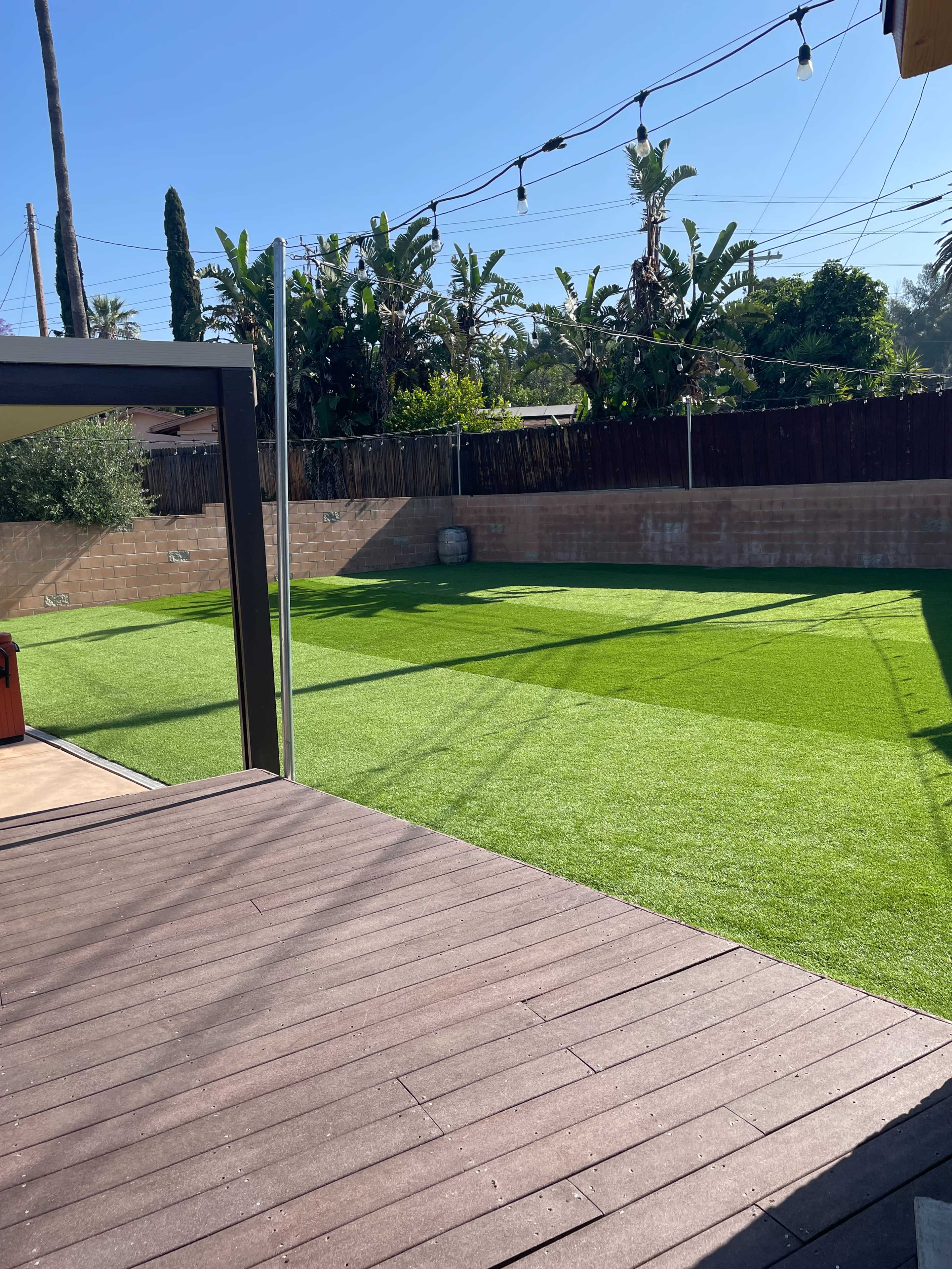 A backyard with artificial grass, framed by a wooden deck and palm trees in the background under a clear blue sky.