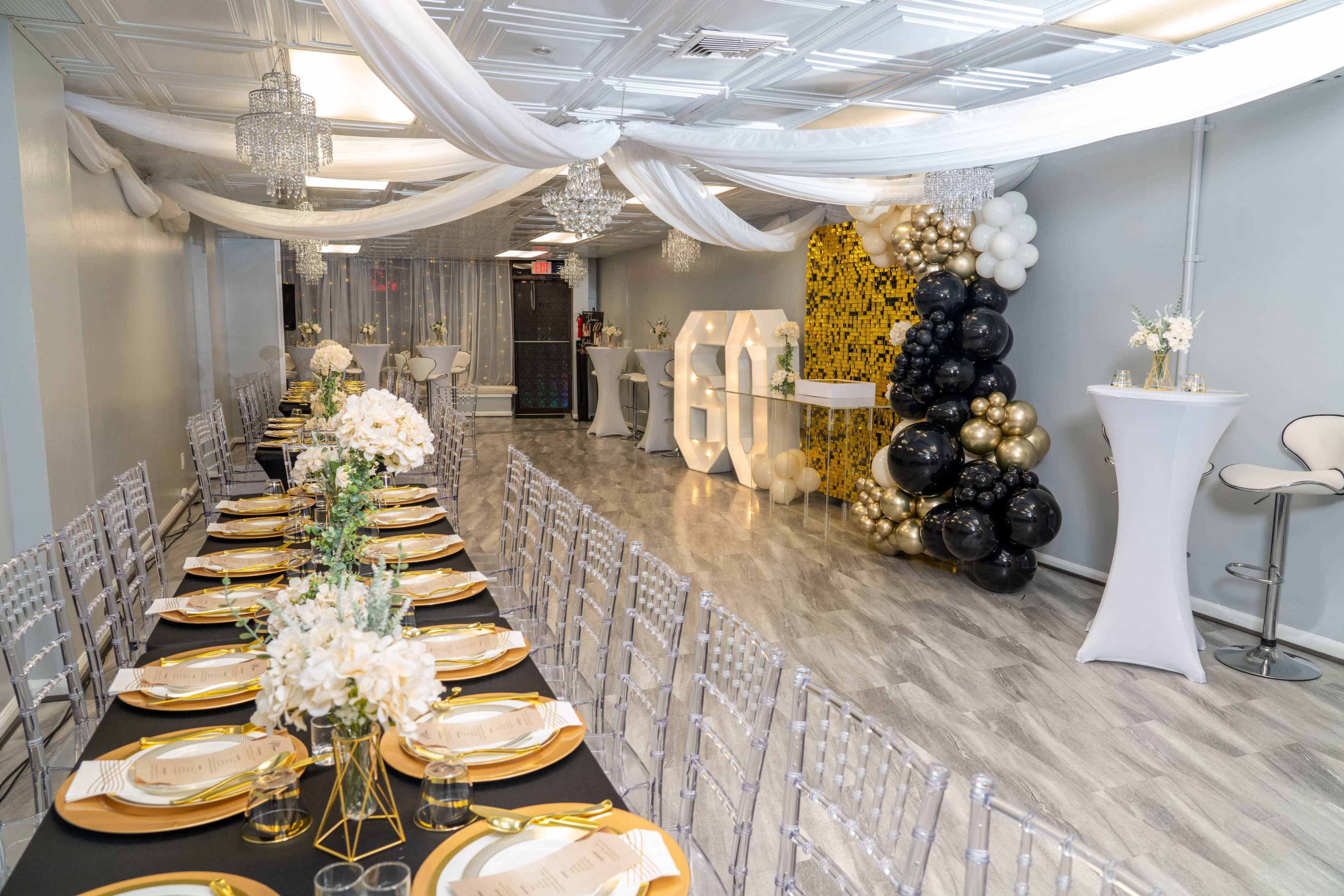 The image shows a decorated banquet hall with a long black and gold dining table set for a celebration, surrounded by flowing white drapery, balloons, and illuminated decorative letters.
