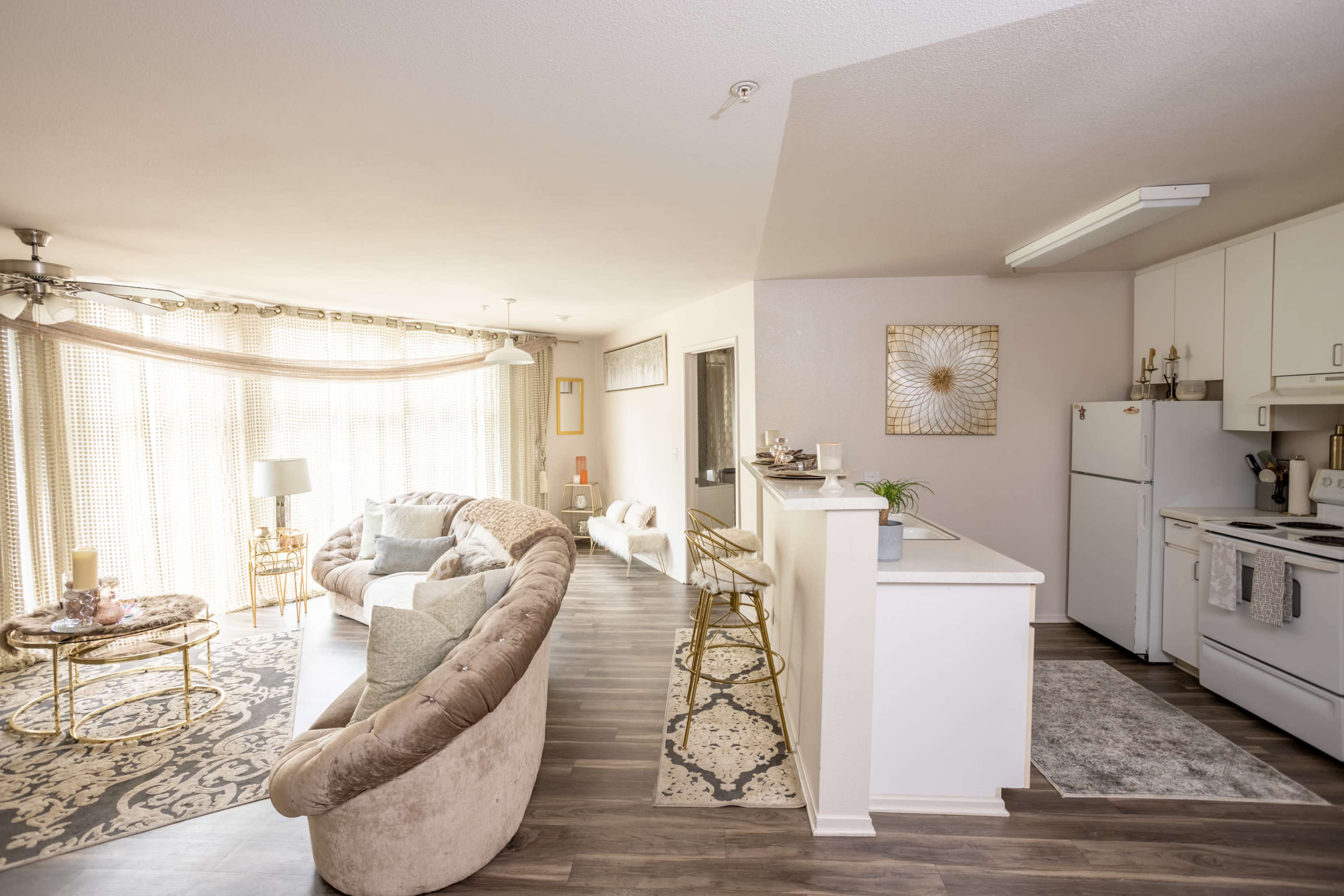 A cozy, open-concept living space features a seating area with a large, curved sofa, a kitchen bar with stools, and large windows letting in natural light.