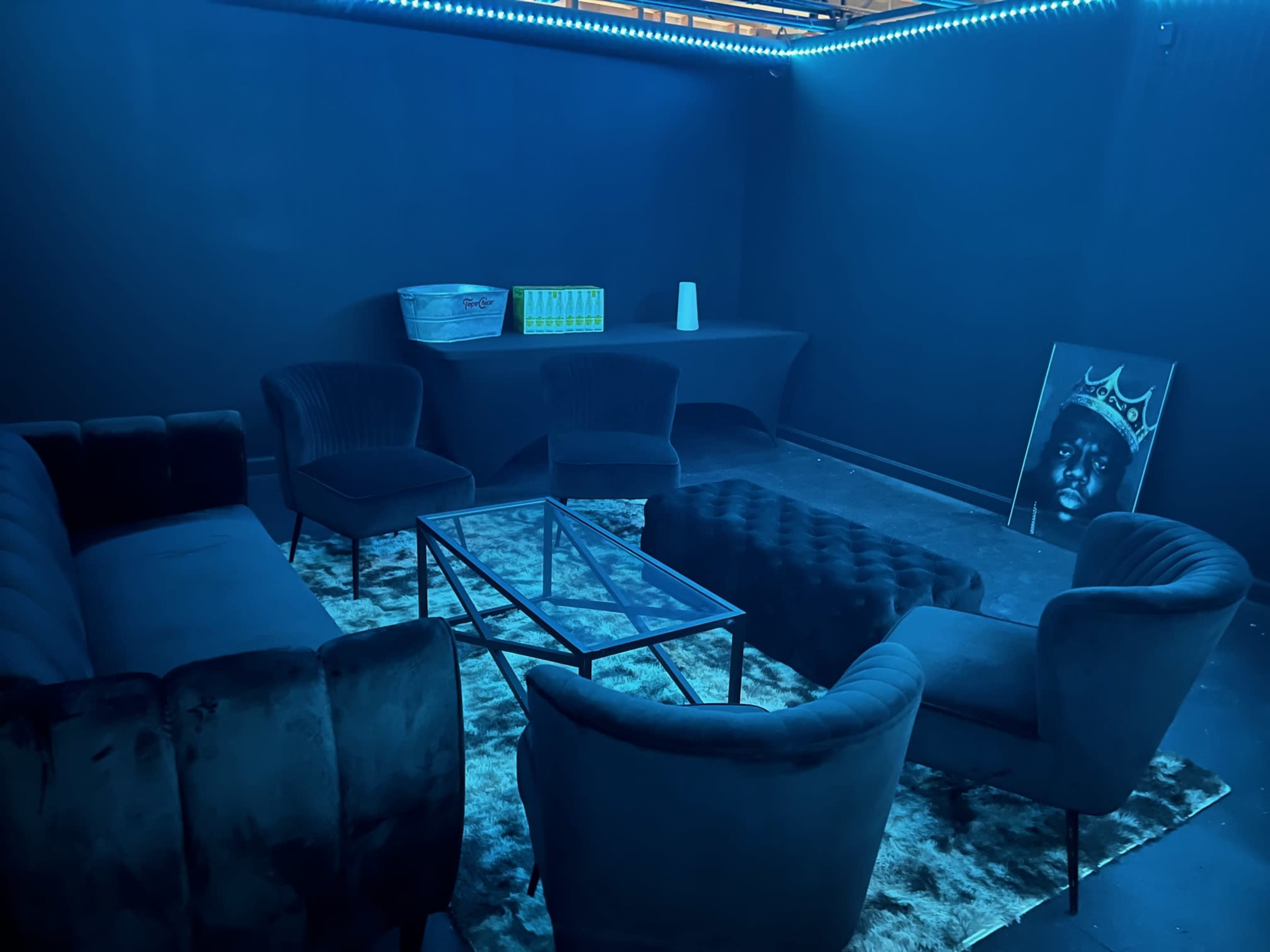 A dimly lit room with blue walls, featuring a black velvet sofa, several armchairs, a glass coffee table, and a framed portrait on the floor.