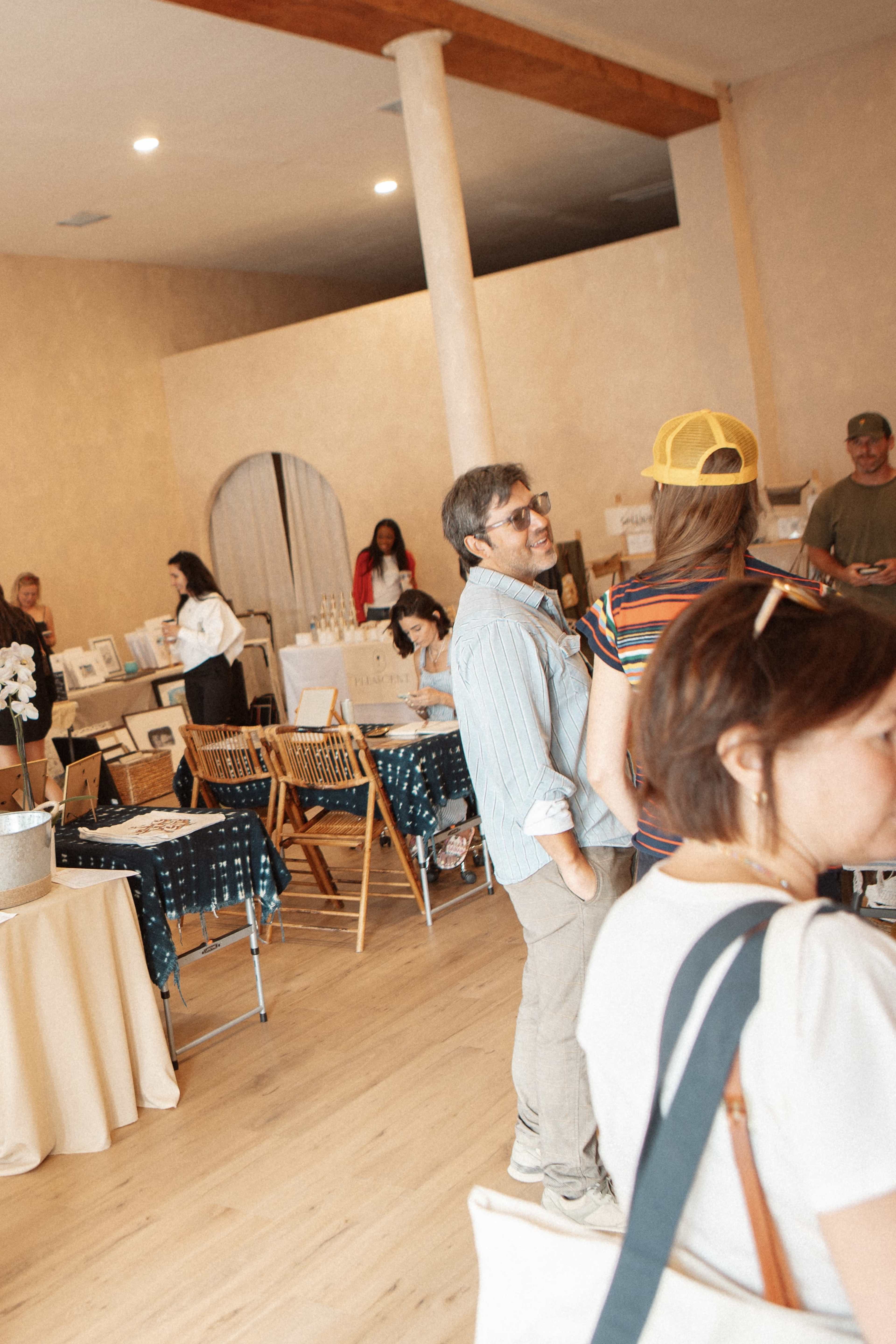 The image shows a gathering of people in a light-colored room featuring several tables displaying various items, with individuals engaged in conversation.
