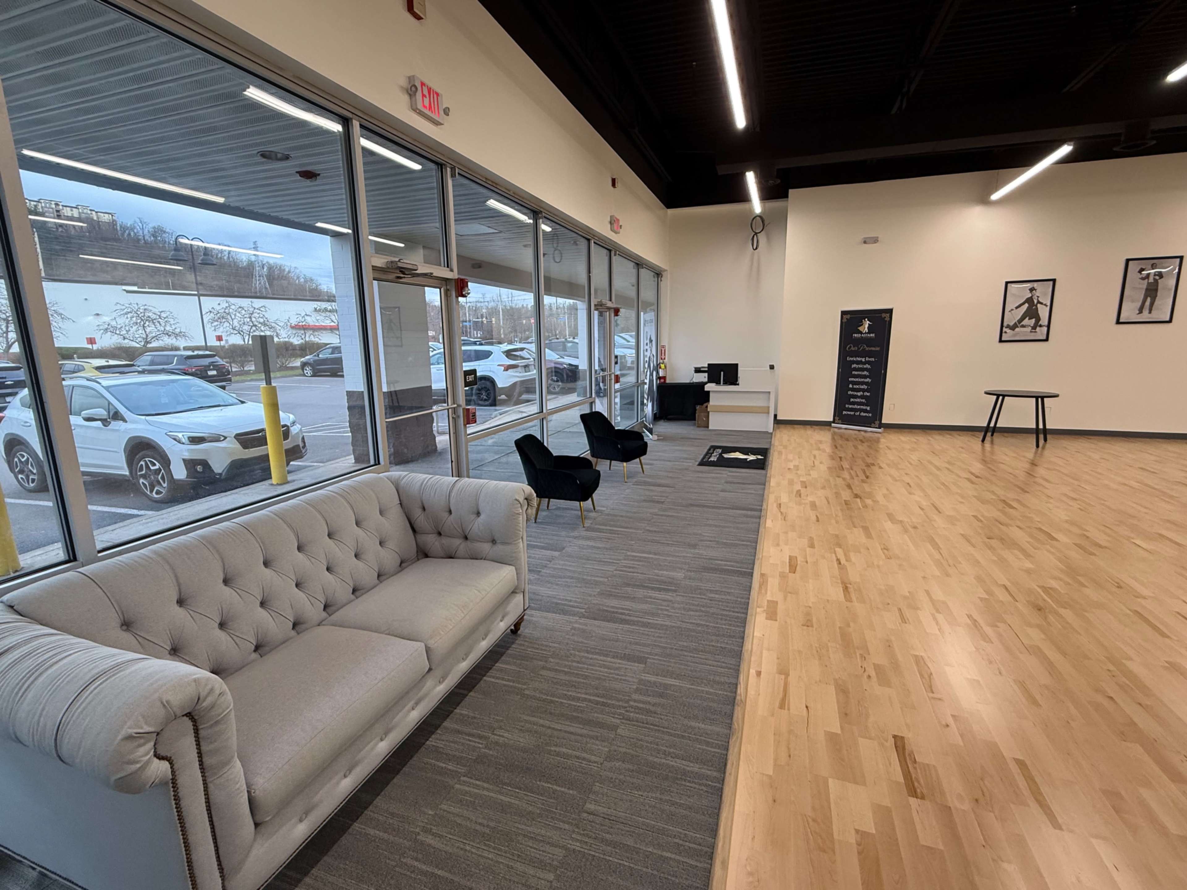 The image shows a modern indoor space featuring a tufted sofa, black chairs, wooden flooring, and large glass windows with a view of parked cars outside.