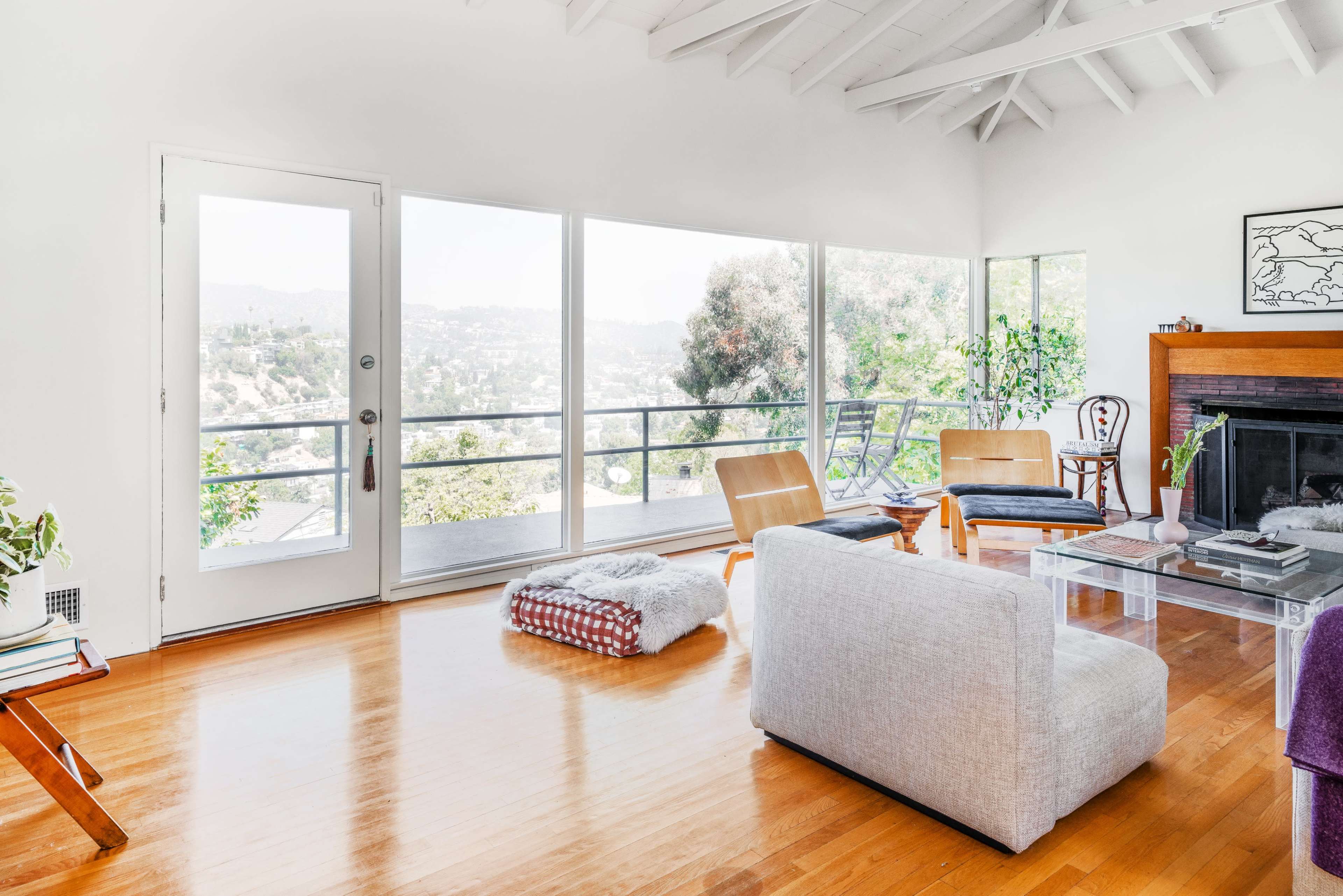 A spacious, bright living room featuring large windows with a view, wooden floors, and minimalist furniture.