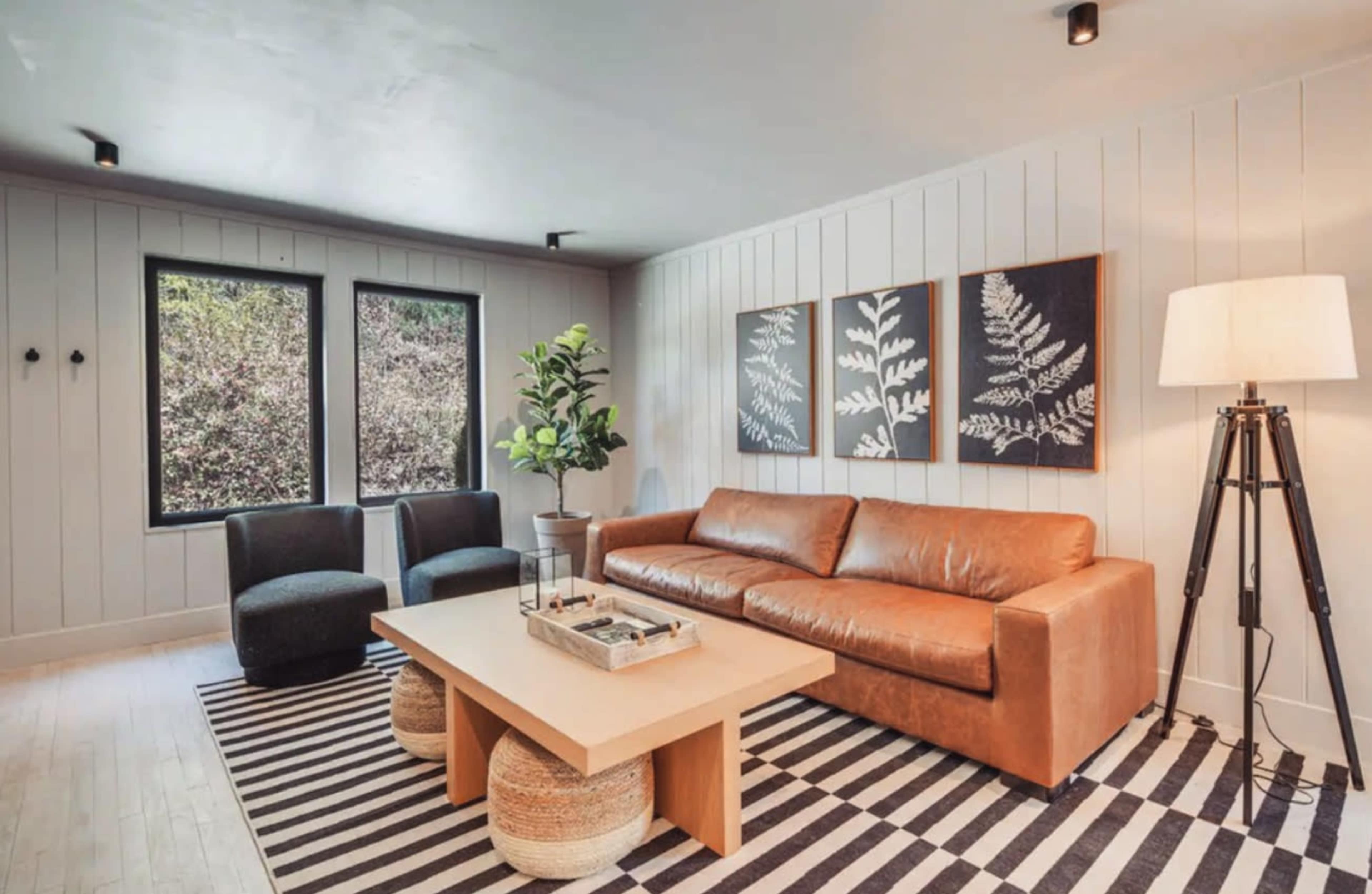 The image shows a modern living room with a brown leather couch, a coffee table, two accent chairs, and framed wall art, set against a backdrop of large windows.
