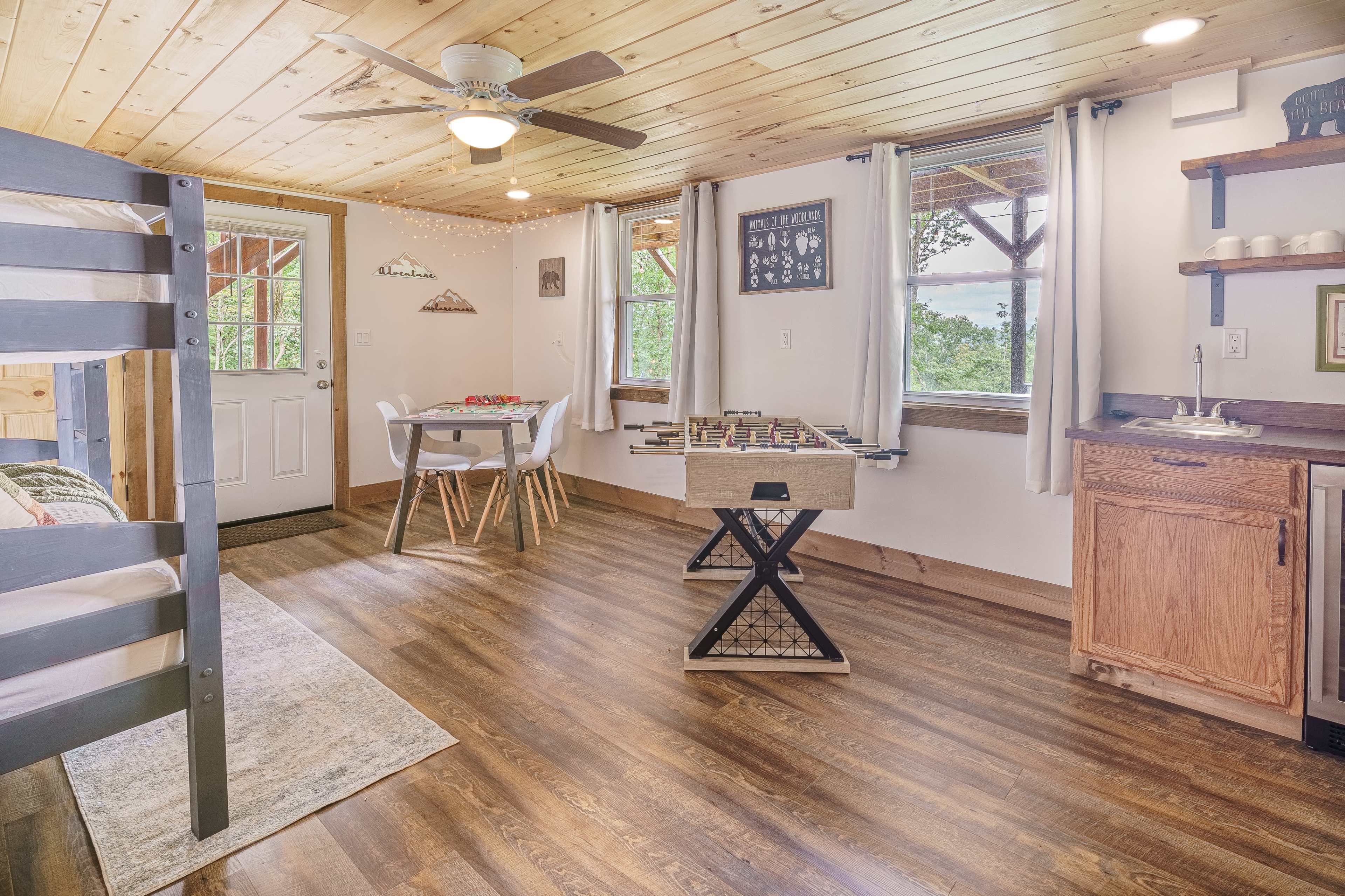 The image shows a cozy interior space with a bunk bed, a dining table, a foosball table, and large windows allowing natural light.