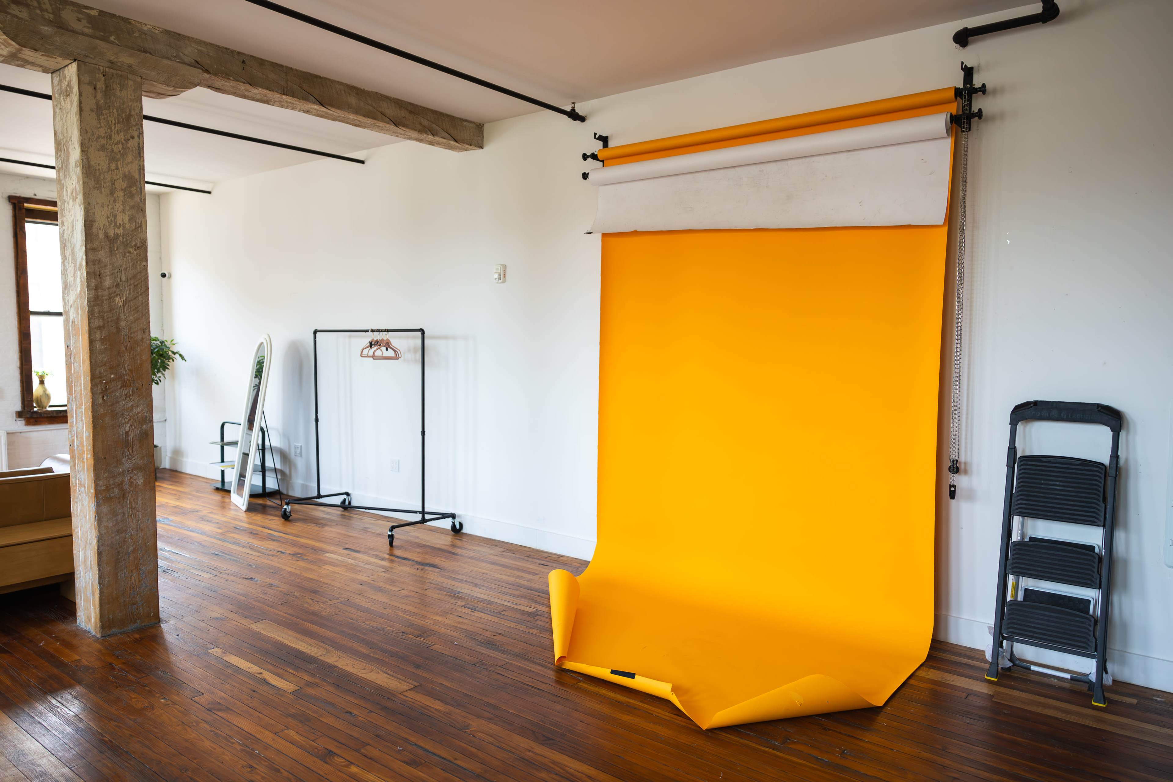 A bright orange backdrop set up in a spacious, minimally furnished room with wooden floors and a few pieces of standing furniture.