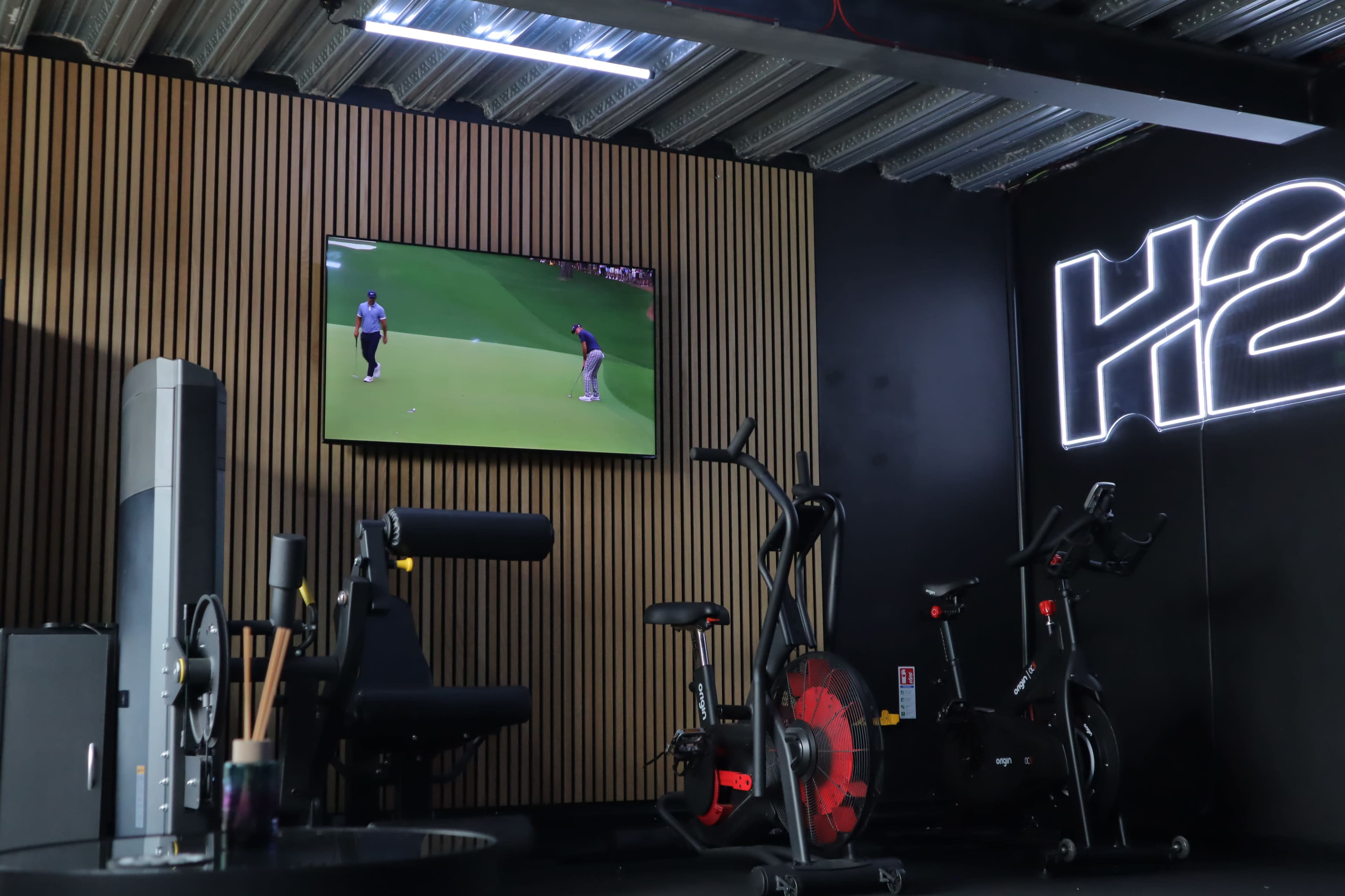 The image shows a gym with exercise equipment, including stationary bikes and strength training machines, along with a television displaying a golf game.