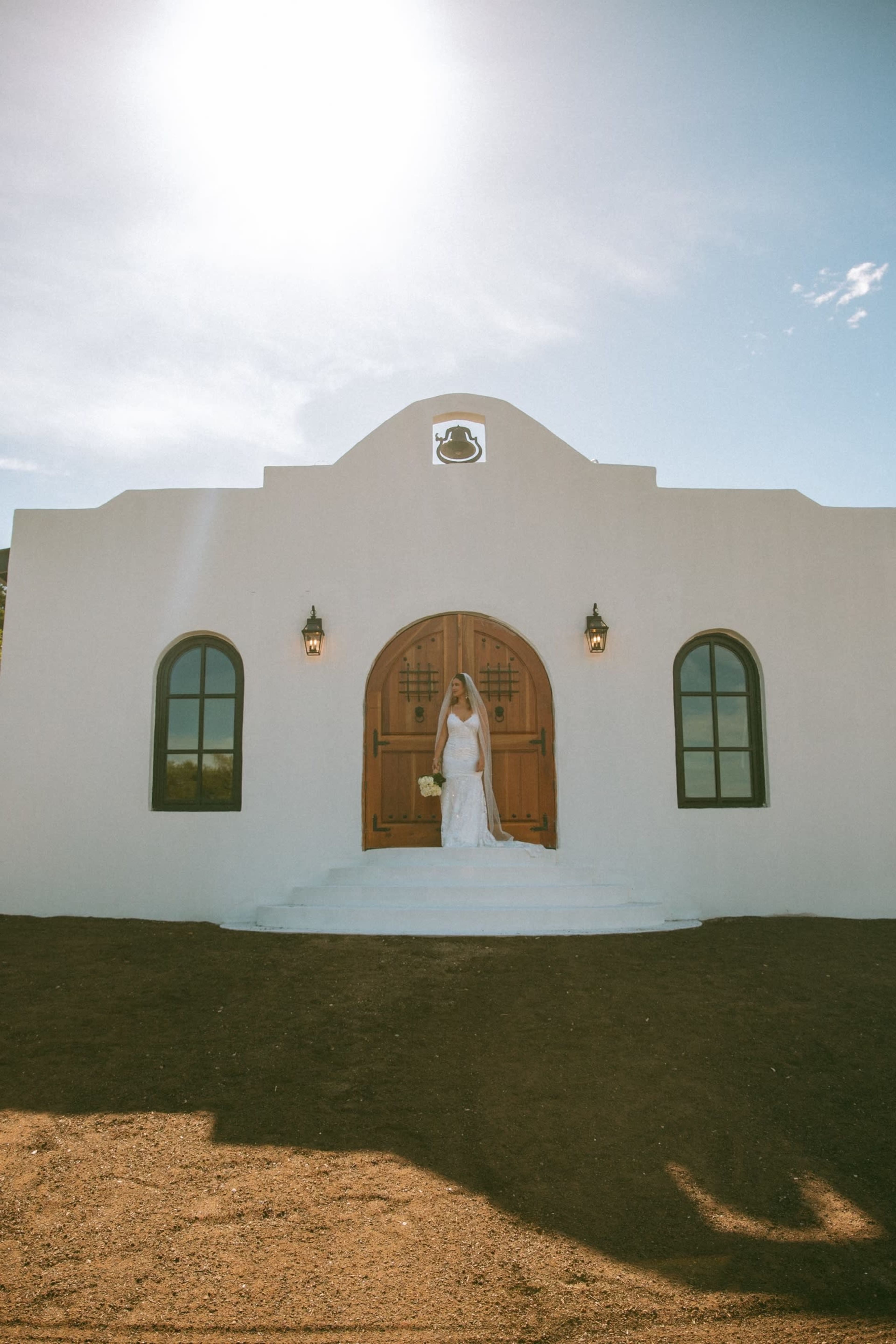 A bride in a white gown stands at the entrance of a white stucco building with wooden doors and large windows.