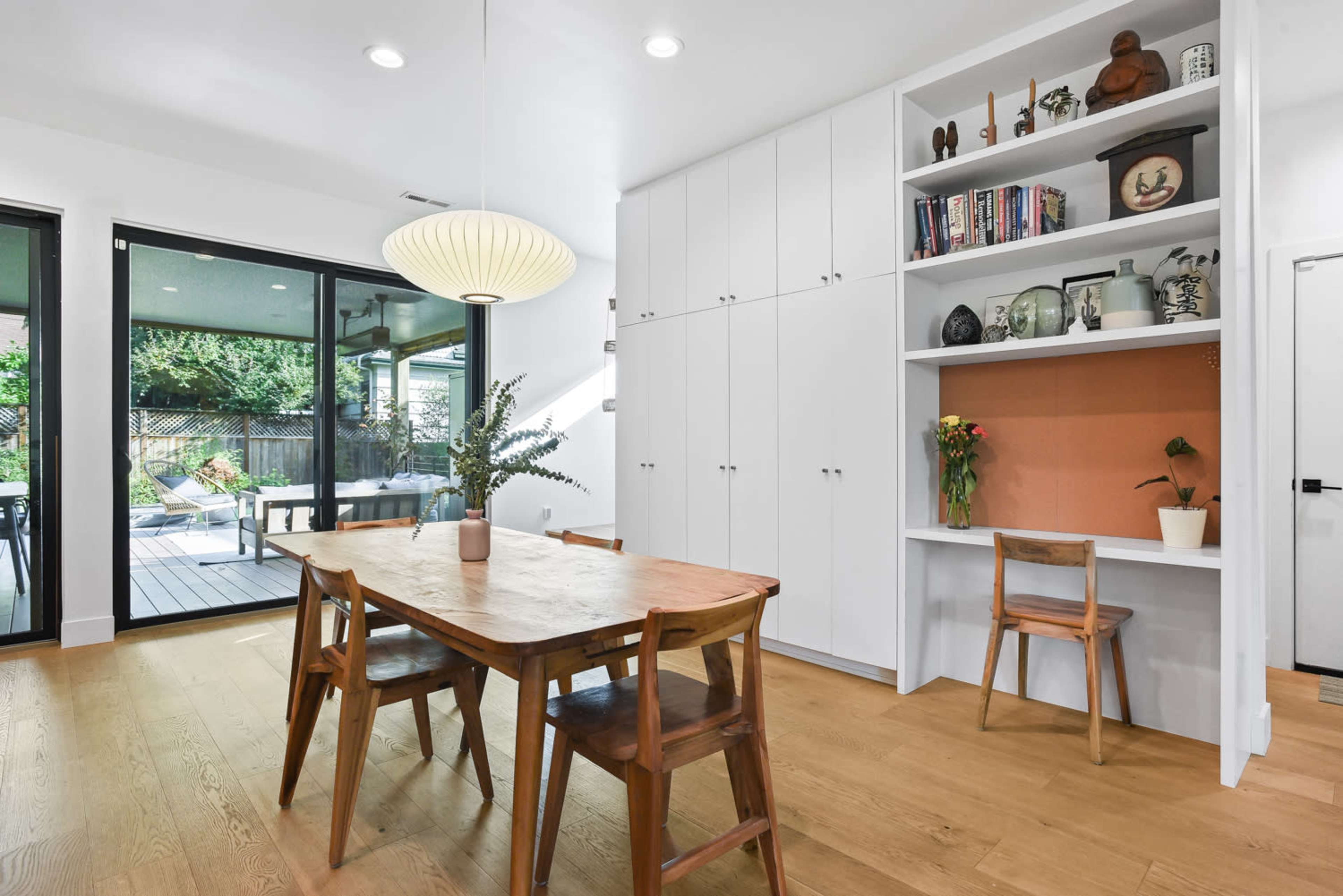 A dining area features a wooden table and chairs, with a built-in shelving unit displaying decorative items, and large windows that open to an outdoor space.